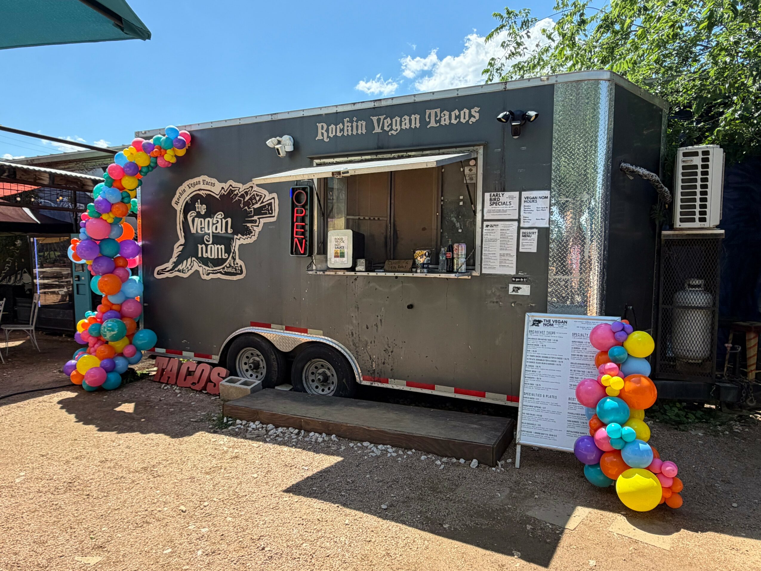 Food truck with colorful balloon decorations on both sides, serving vegan tacos, parked outdoors with trees in background.