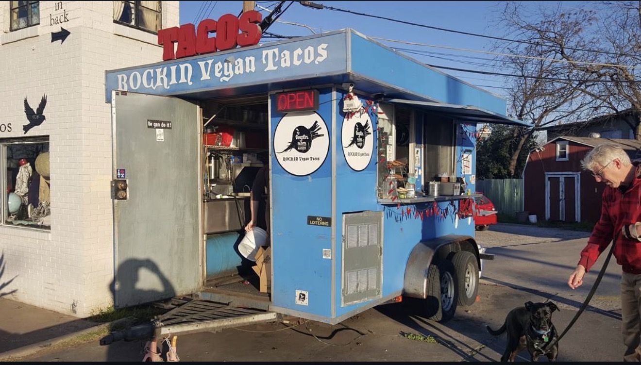 Vegan taco stand with blue and white exterior, sign reading 'Rockin Vegan Tacos,' and a person walking a dog nearby.