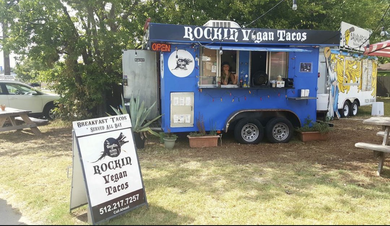 Blue food truck with signboard outside, surrounded by trees and picnic tables, serving vegan tacos.