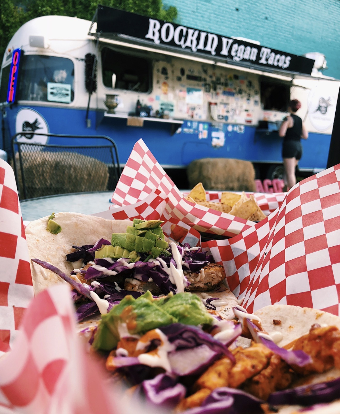 Food on a table with red checkered paper, with a food truck and person in background, outdoor setting.