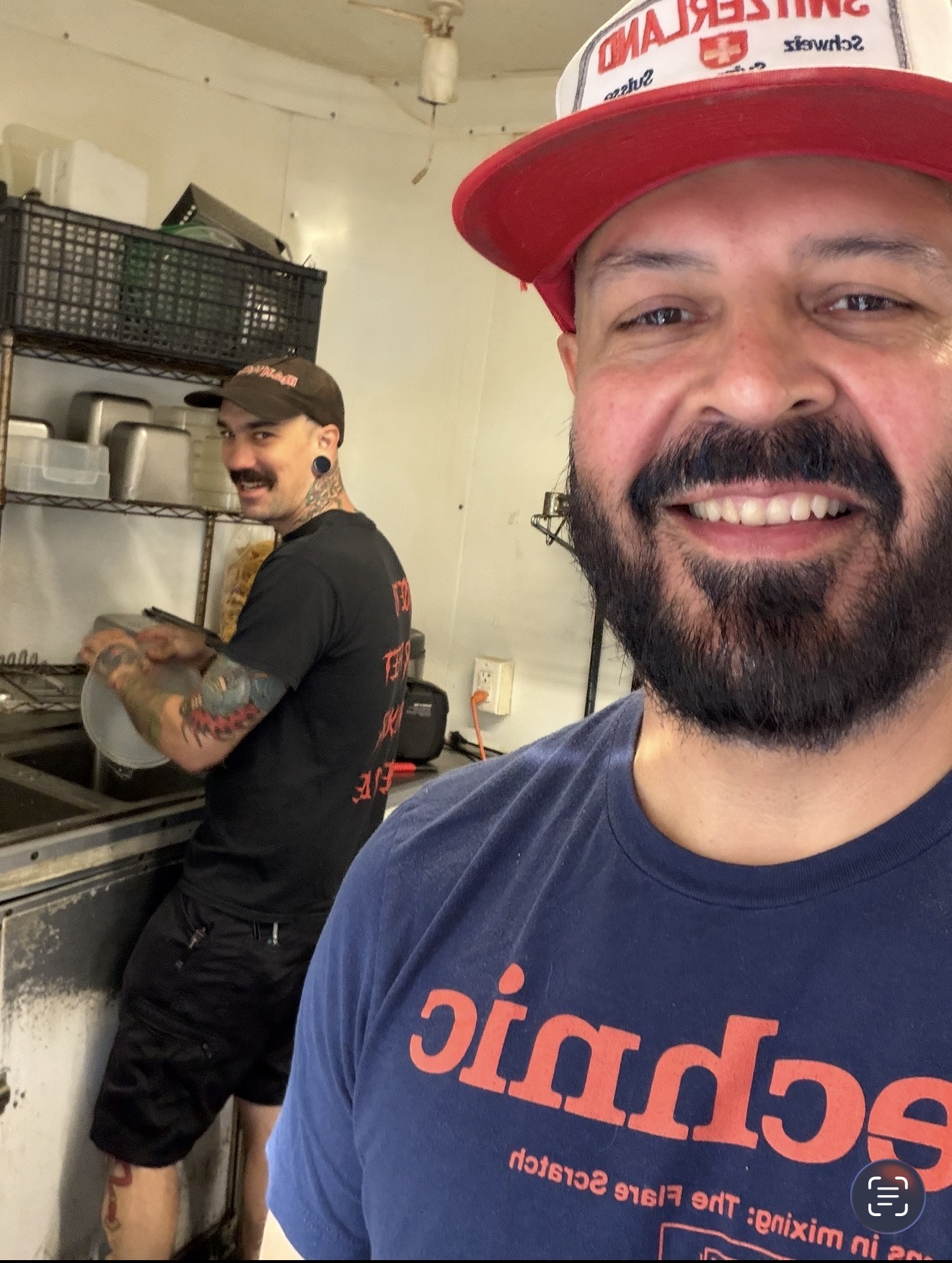 Two men in a kitchen, one smiling in the foreground wearing a red cap and blue shirt, the other cooking in the background.