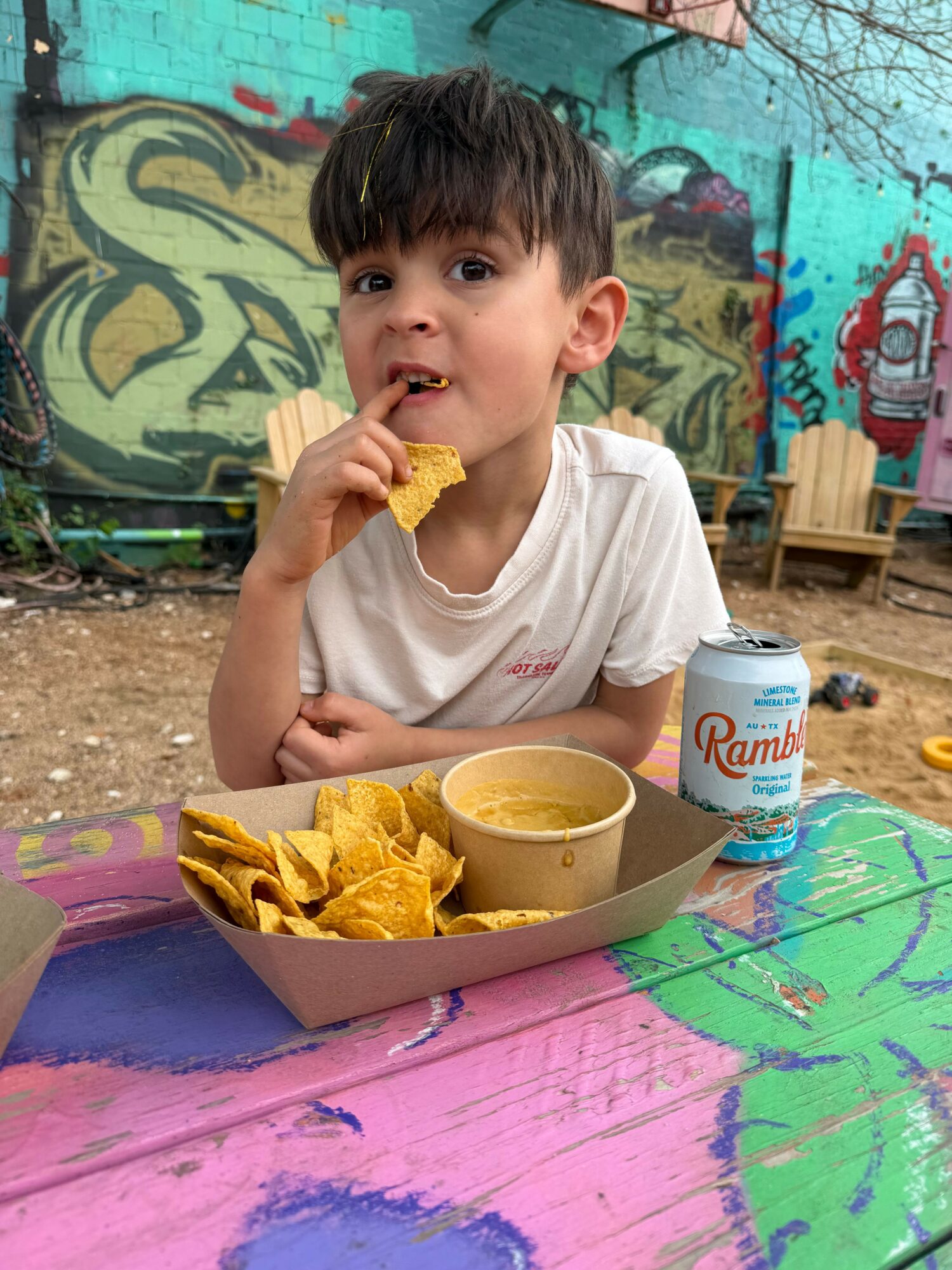 Child eating chips at colorful table outdoors, graffiti background, with a drink and a bowl of chips.
