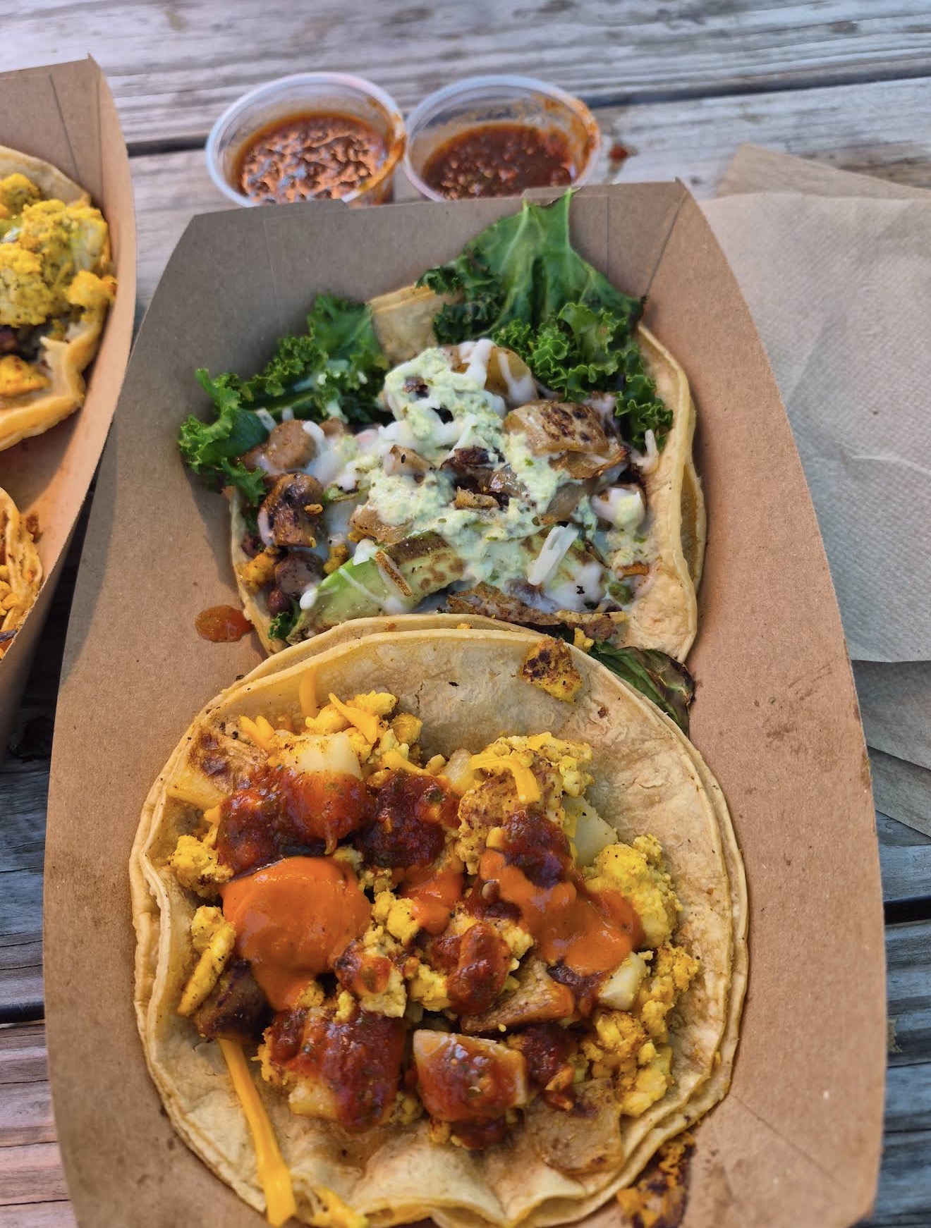 Two tacos with meat, vegetables, and cheese on a paper-lined tray, with two small cups of salsa in the background.
