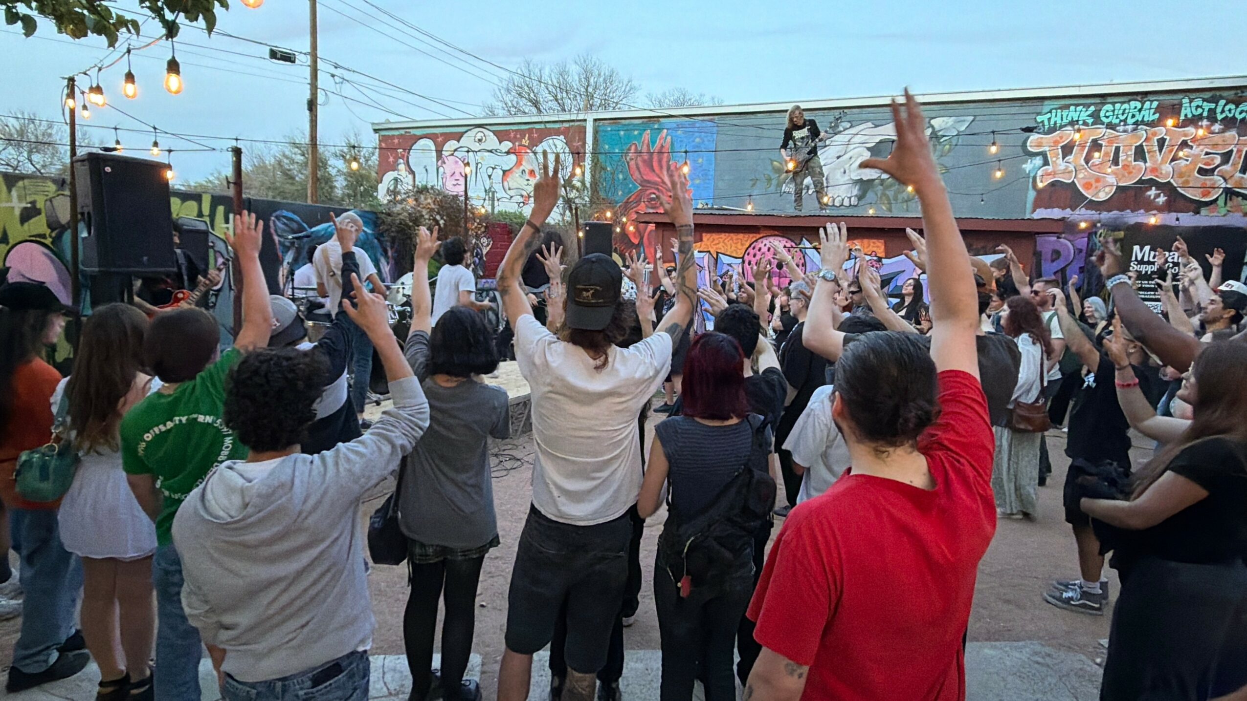 Crowd of people with raised hands watching a performer on an outdoor stage, graffiti art background, evening setting.