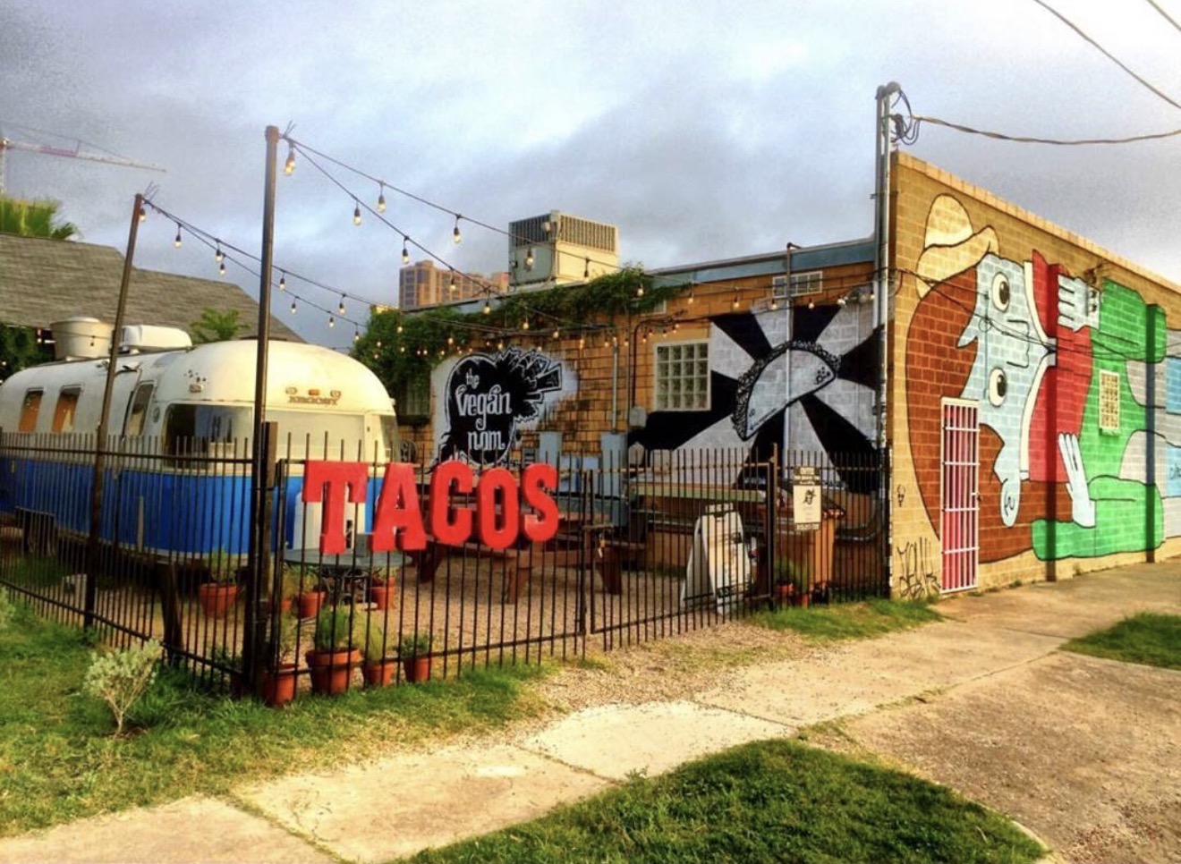 Colorful mural on brick building with a train and a fence in front, featuring a sign that says 'TACOS'.