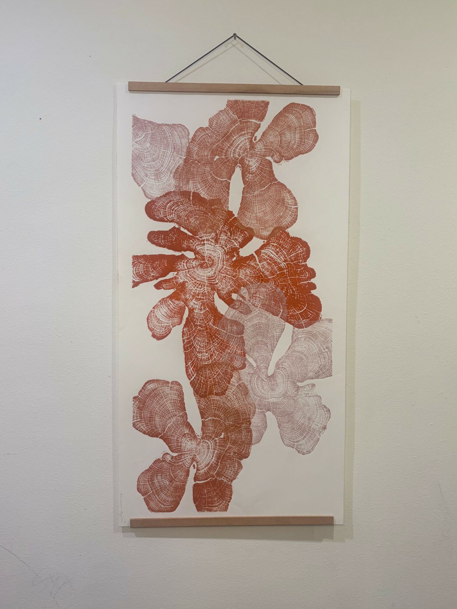Vertical hanging artwork with abstract reddish-brown shapes on a white background, mounted on a wall with wooden rods at top and bottom.