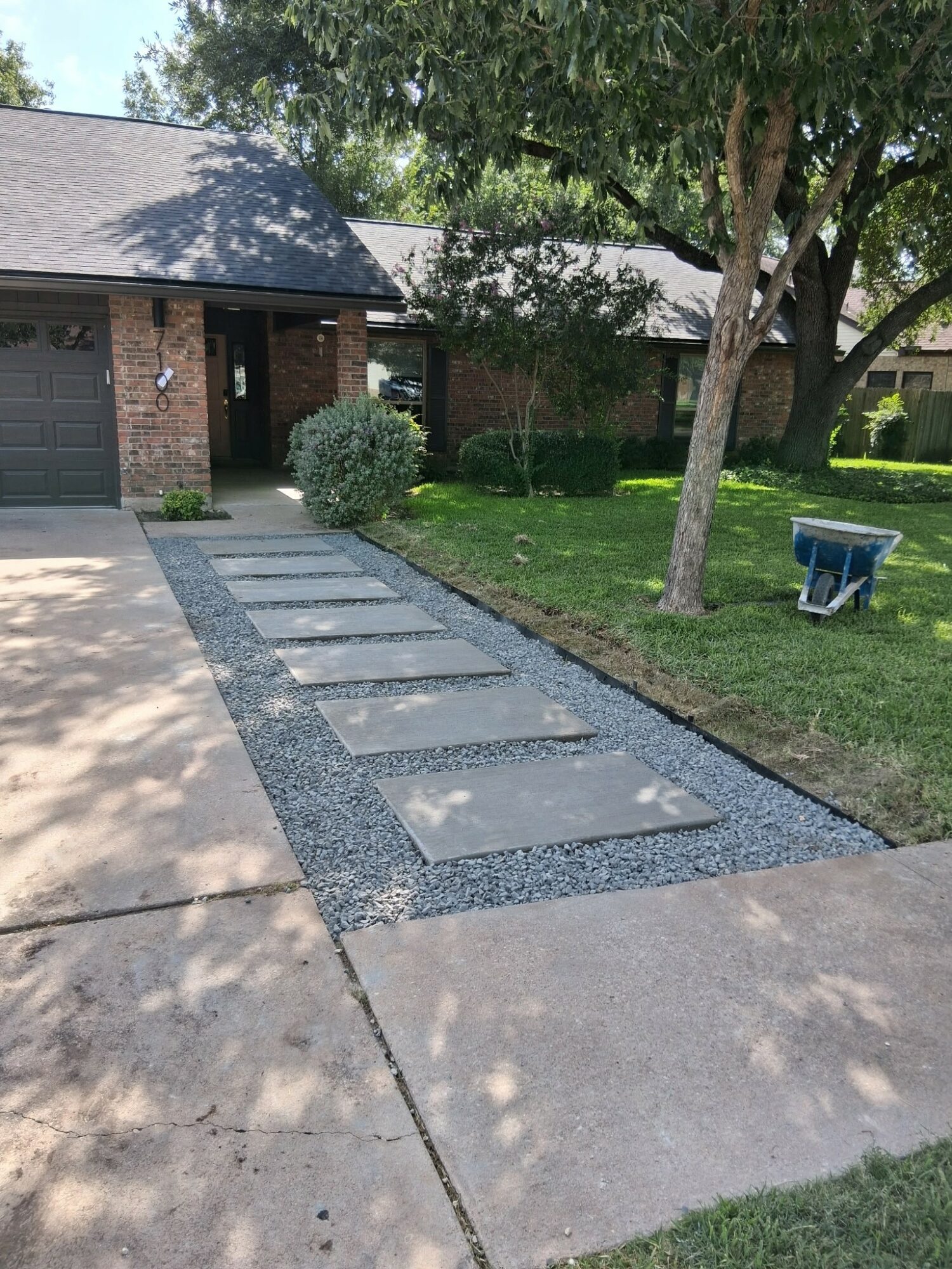 Concrete pathway with large square stepping stones leading to a house, surrounded by grass and trees, with a wheelbarrow nearby.