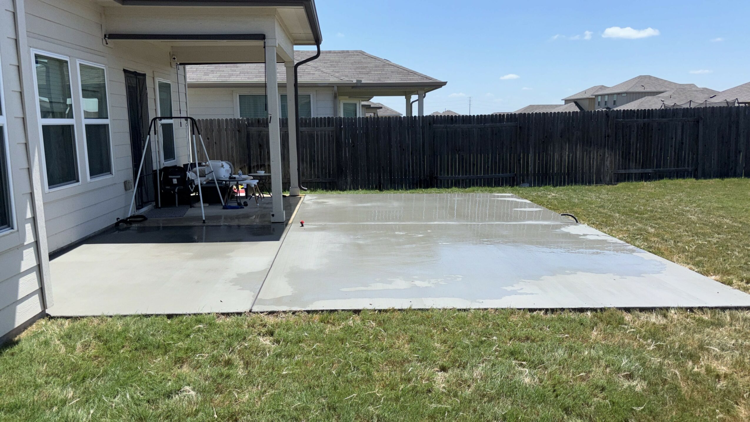 Concrete patio extending from house with a grassy yard, black fence, and neighboring houses in background.