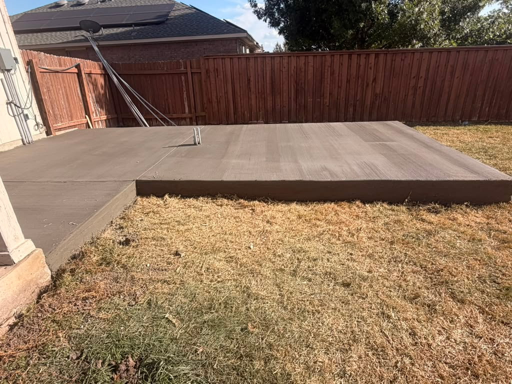 Concrete patio with a raised edge, adjacent to a grassy yard, enclosed by a wooden fence, with a clothesline in the background.