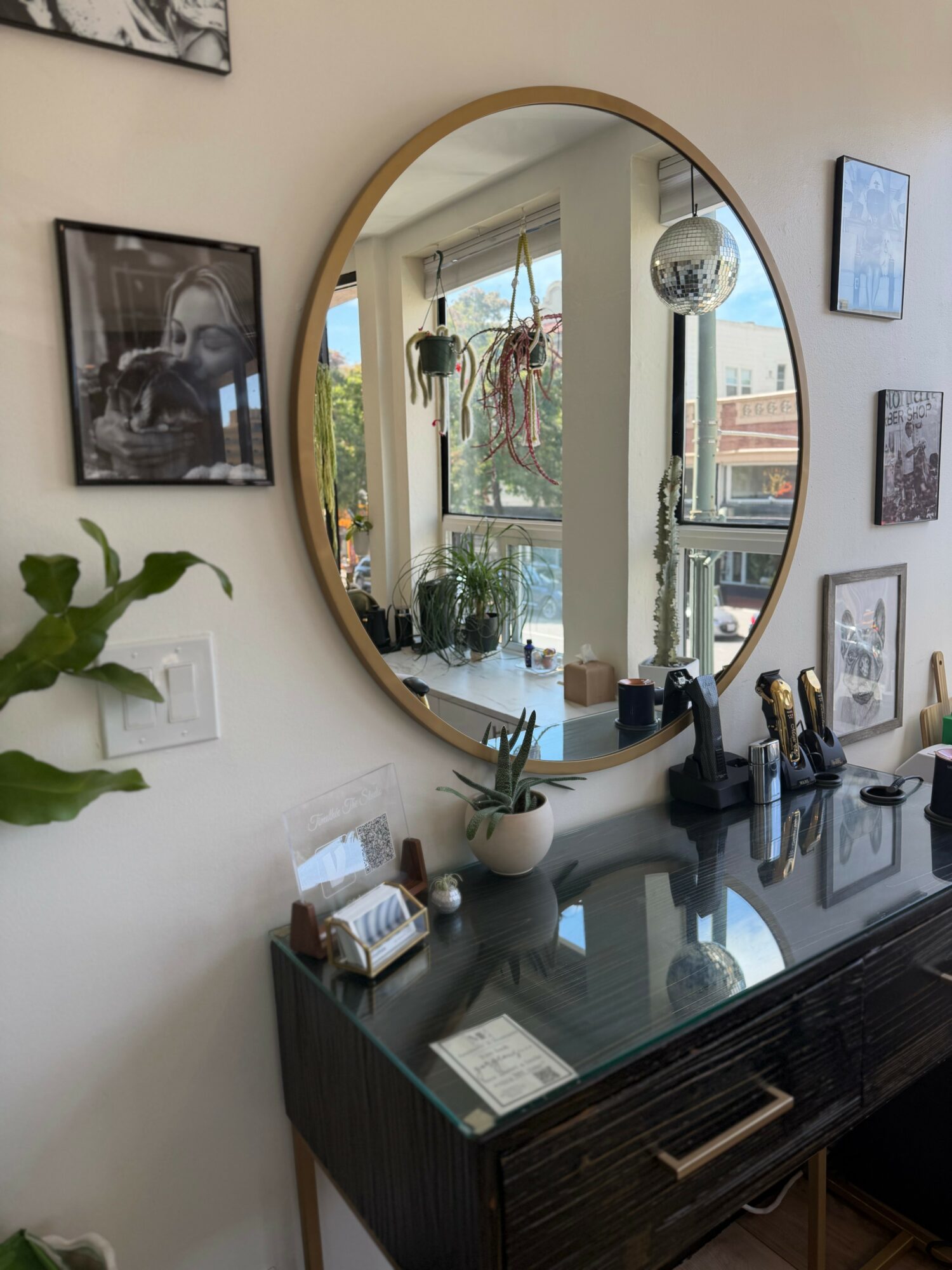 Round mirror reflecting window and plants, black sideboard with decorative items, framed pictures on wall, and a plant.