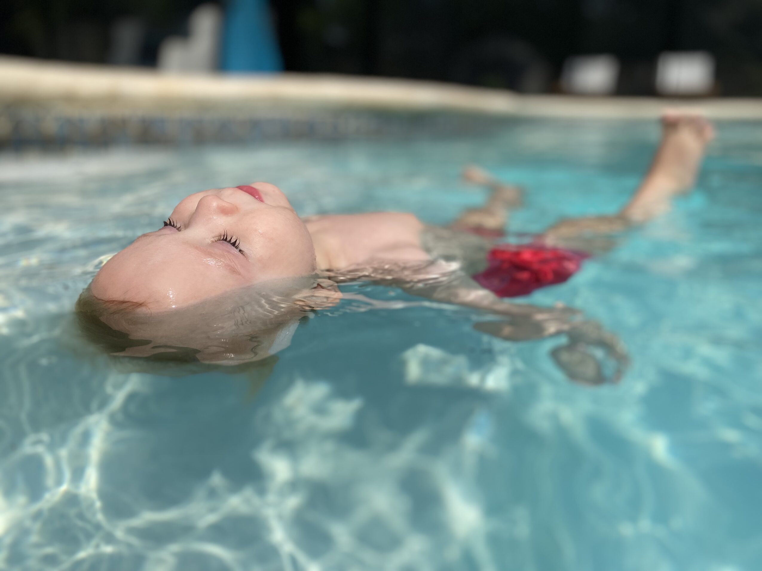 Child floating on back in a swimming pool with eyes closed, wearing red swim trunks, water reflecting sunlight.