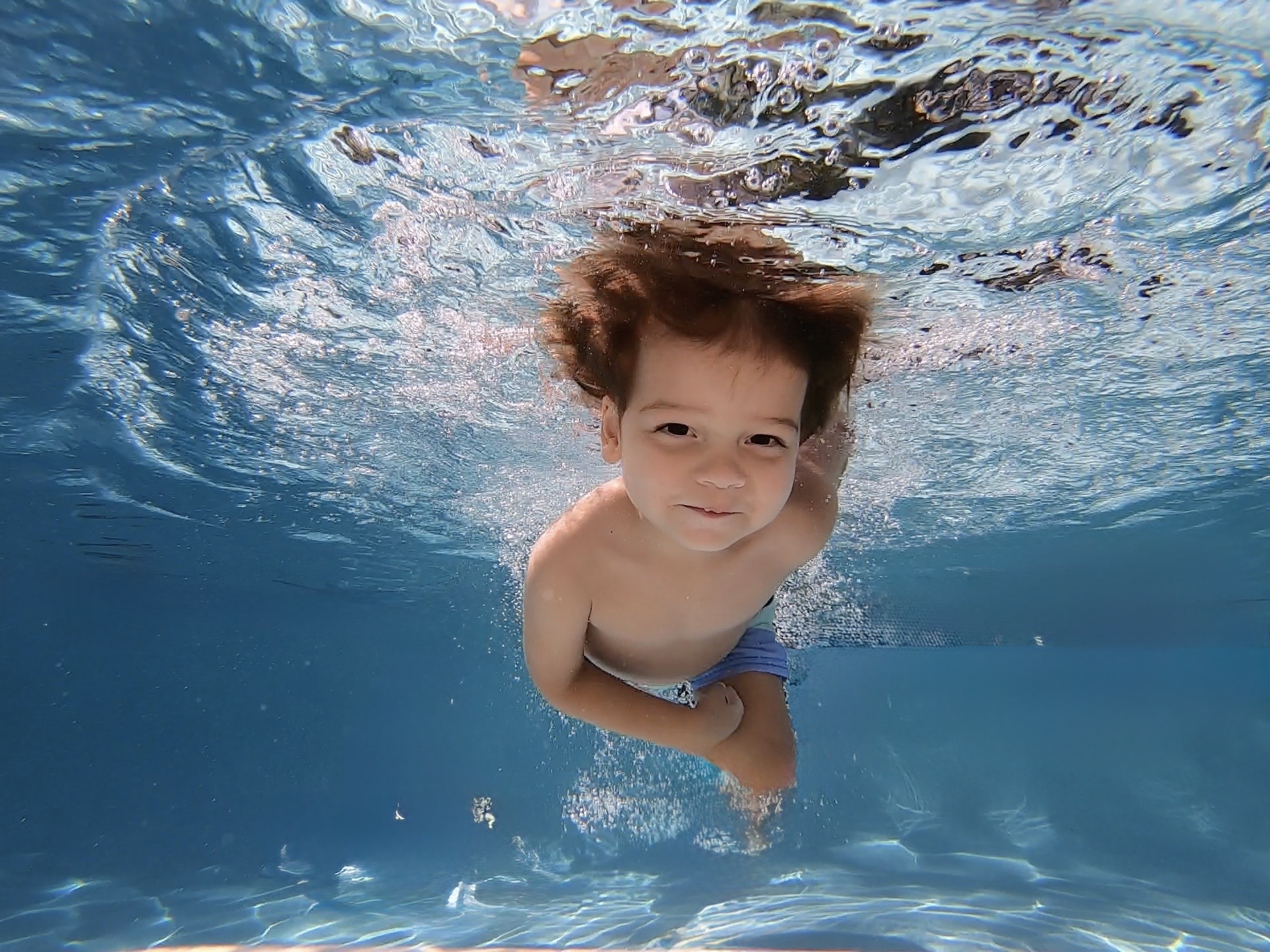 Child swimming underwater, smiling, with hair floating and water ripples around.
