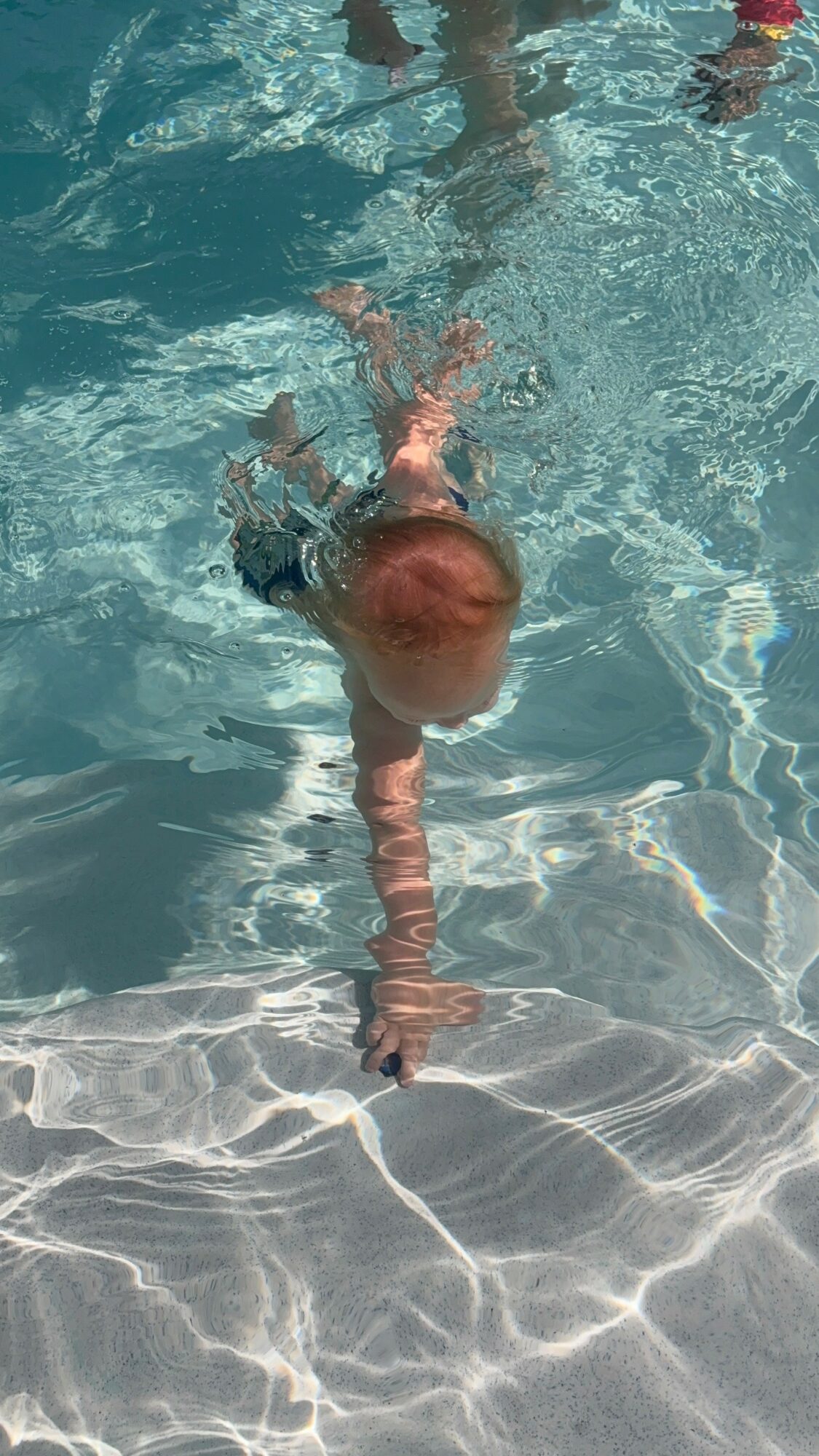 Child swimming underwater in a pool, reaching towards the pool floor with one arm extended.