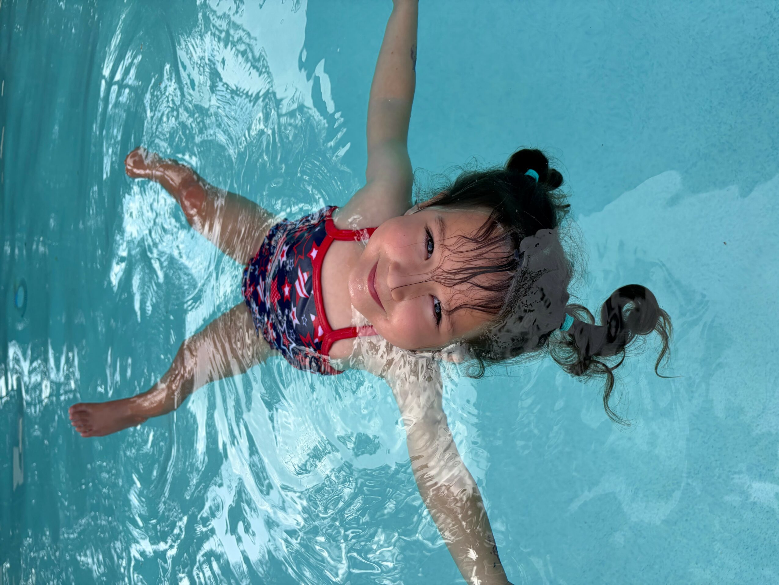 Young girl floating in a swimming pool with arms outstretched, wearing a colorful swimsuit, smiling, with water ripples around her.