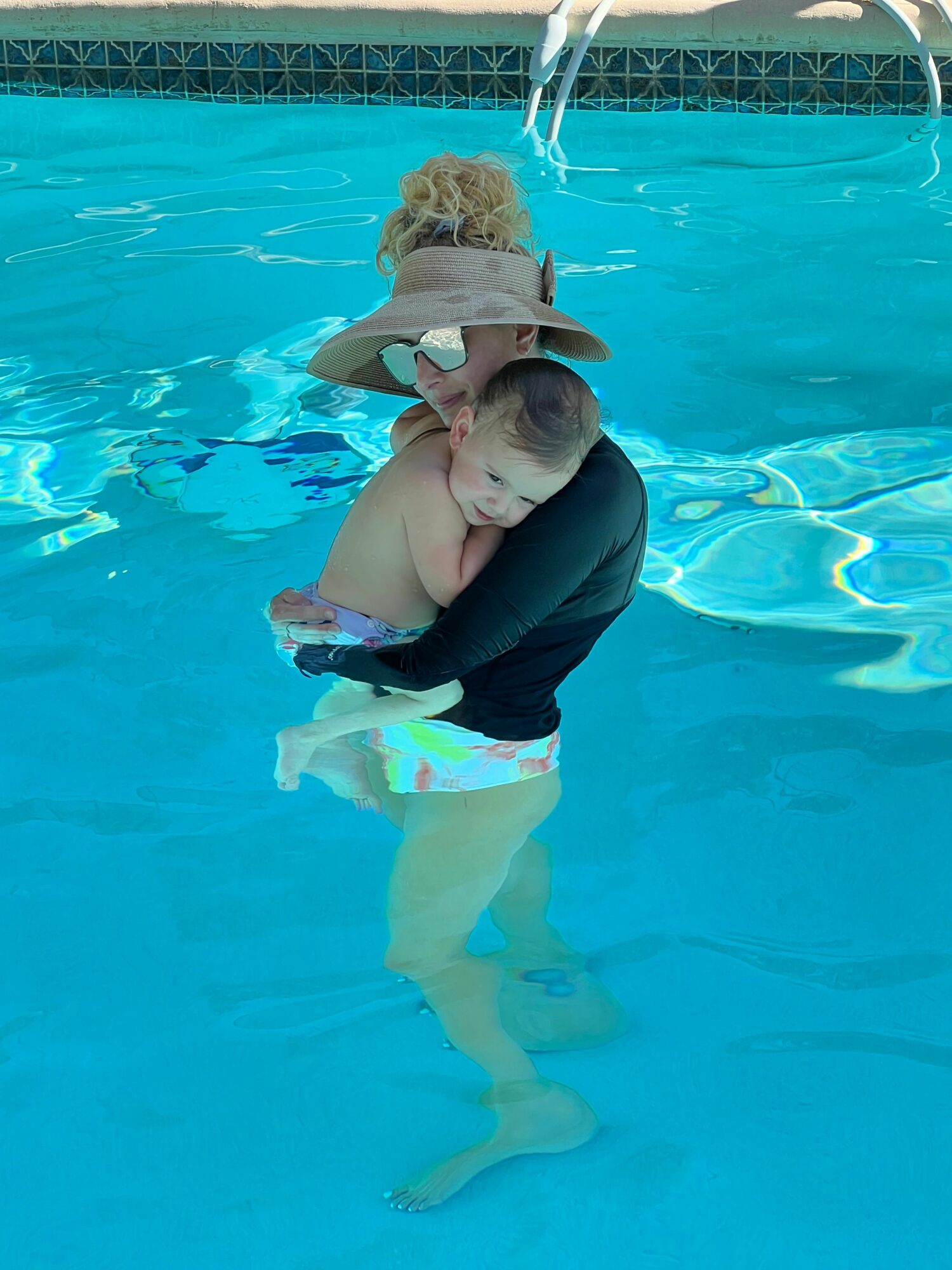 Person holding a child in a swimming pool, child wearing a hat and black clothing, water is blue and clear.