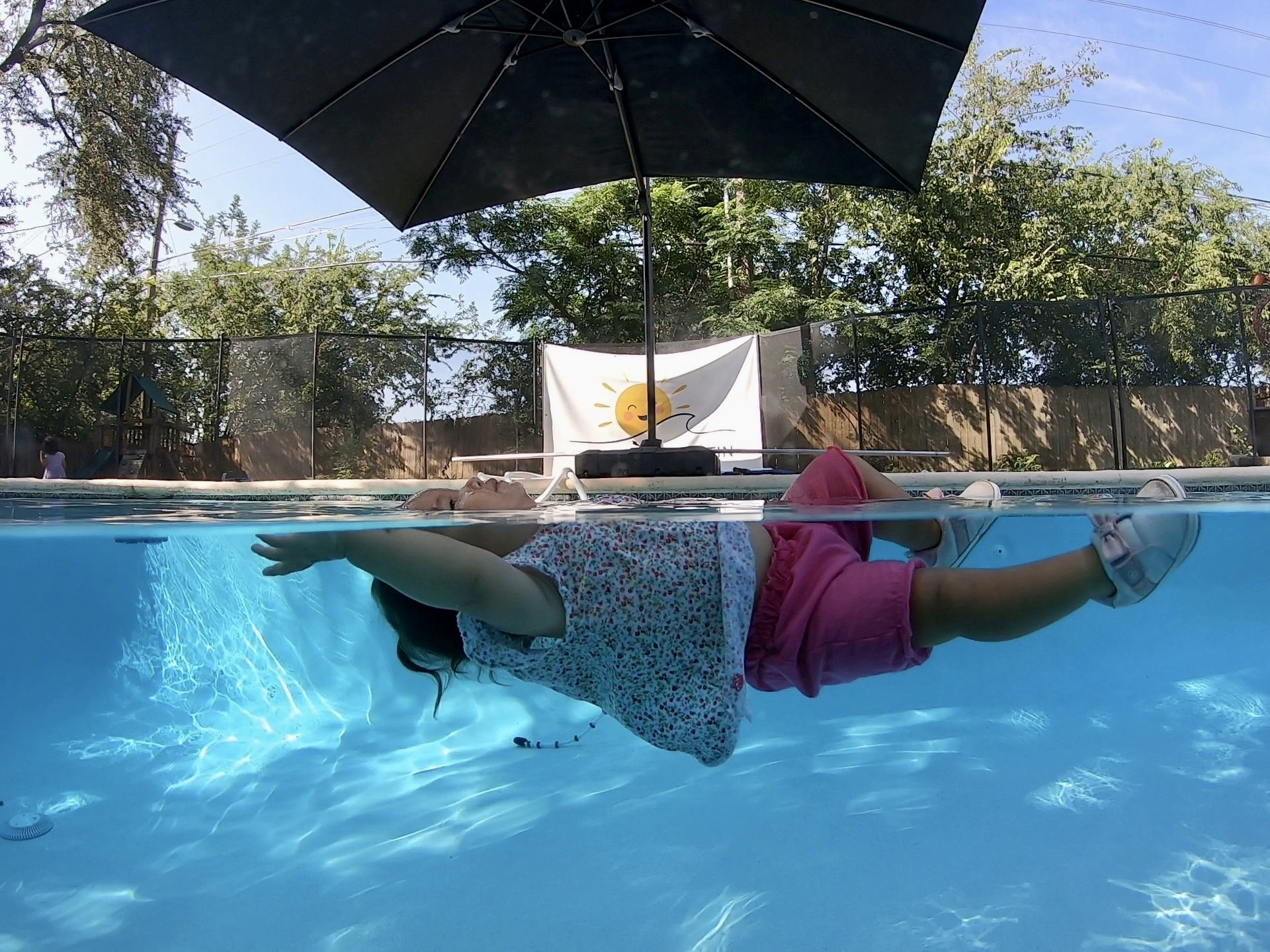 Child swimming underwater in a pool, wearing a pink shirt and shorts, with an umbrella and trees outside the pool. Word count: 20