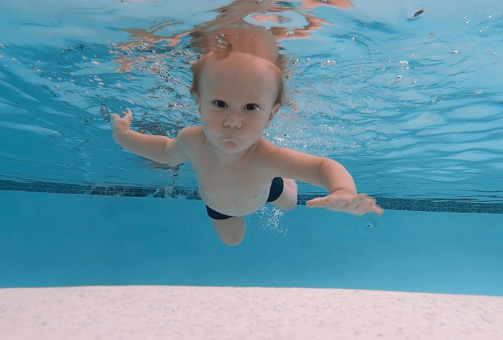 Young child swimming underwater in a pool, looking at the camera with arms extended.