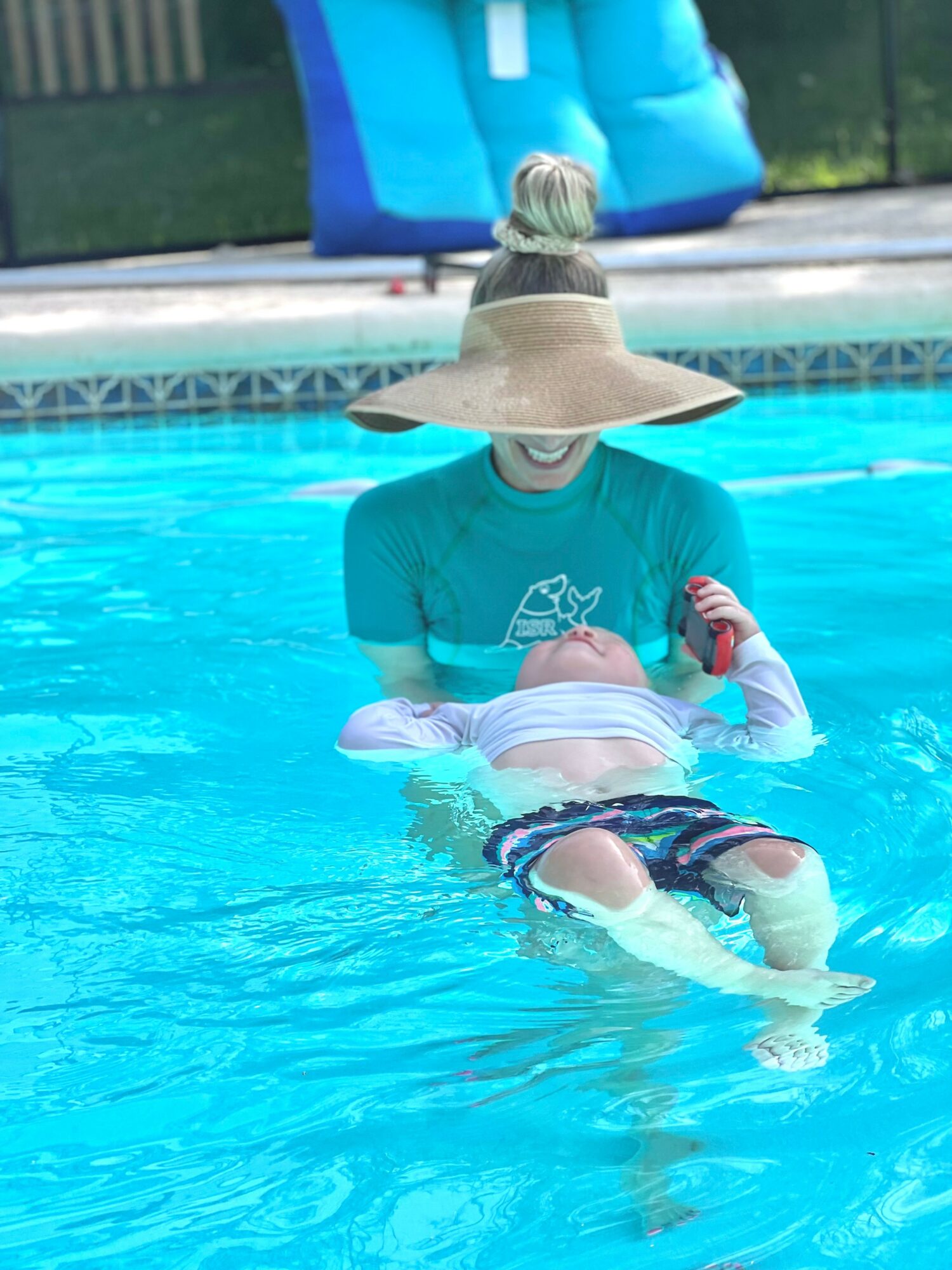 Person in a pool wearing a large sun hat and a teal shirt, holding a child in the water.