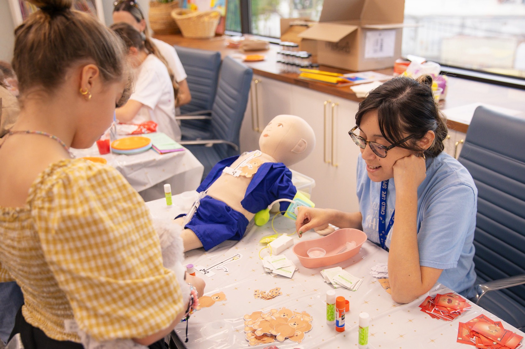 A woman with glasses and a blue shirt sits at a table with a medical mannequin, children, and medical supplies in a room.