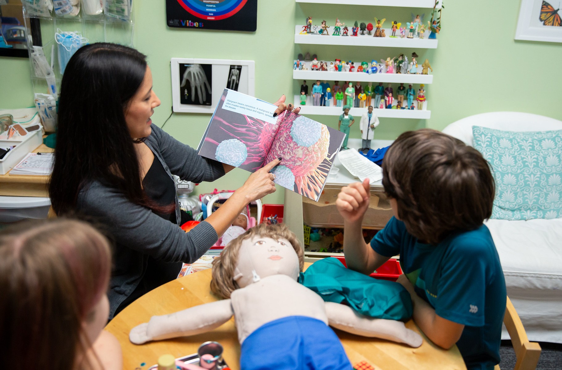 A woman shows a diagram of lungs to children in a medical setting, with a doll on the table and medical equipment nearby.