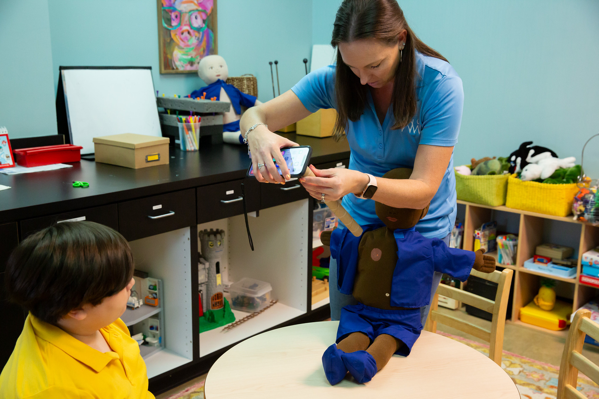 A woman kneels on a table, using a smartphone, with a young child in a yellow shirt nearby in a colorful room.