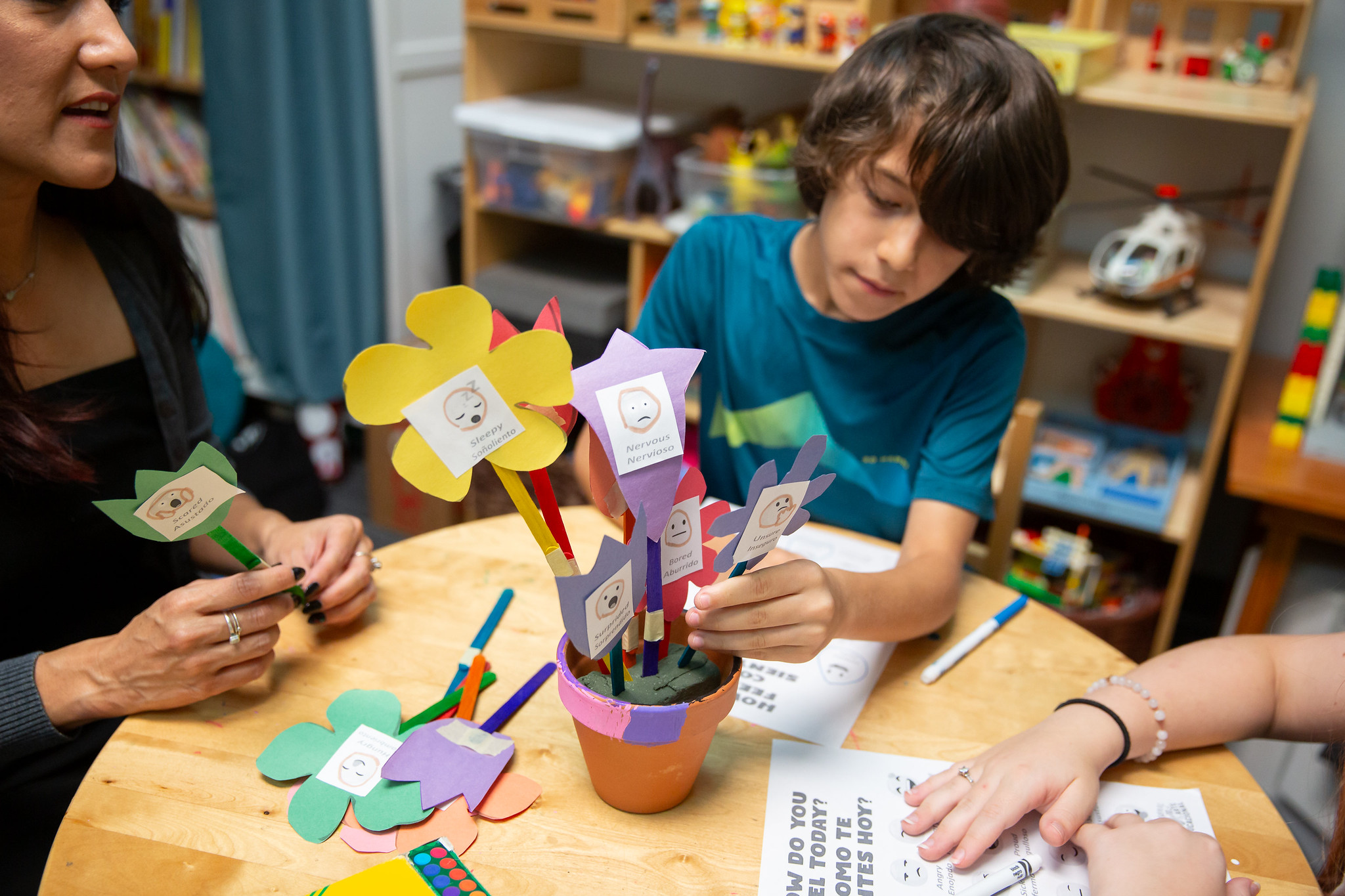 Children and an adult working on paper craft projects at a table in a classroom, with shelves of toys and books in the background.