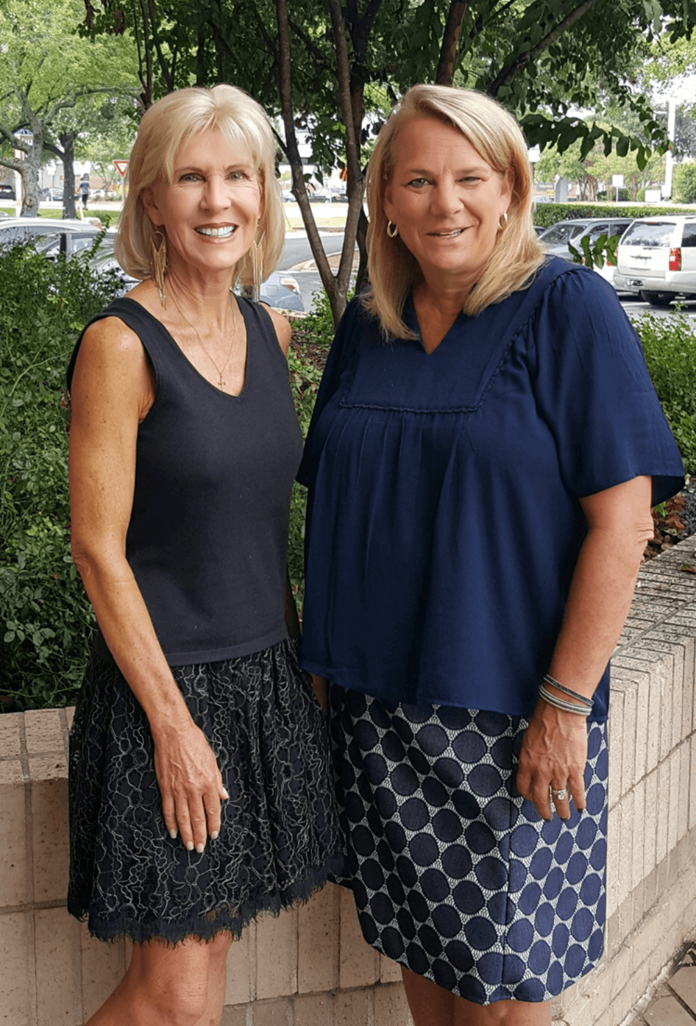 Two women standing outdoors, smiling, with trees and parked cars in the background.