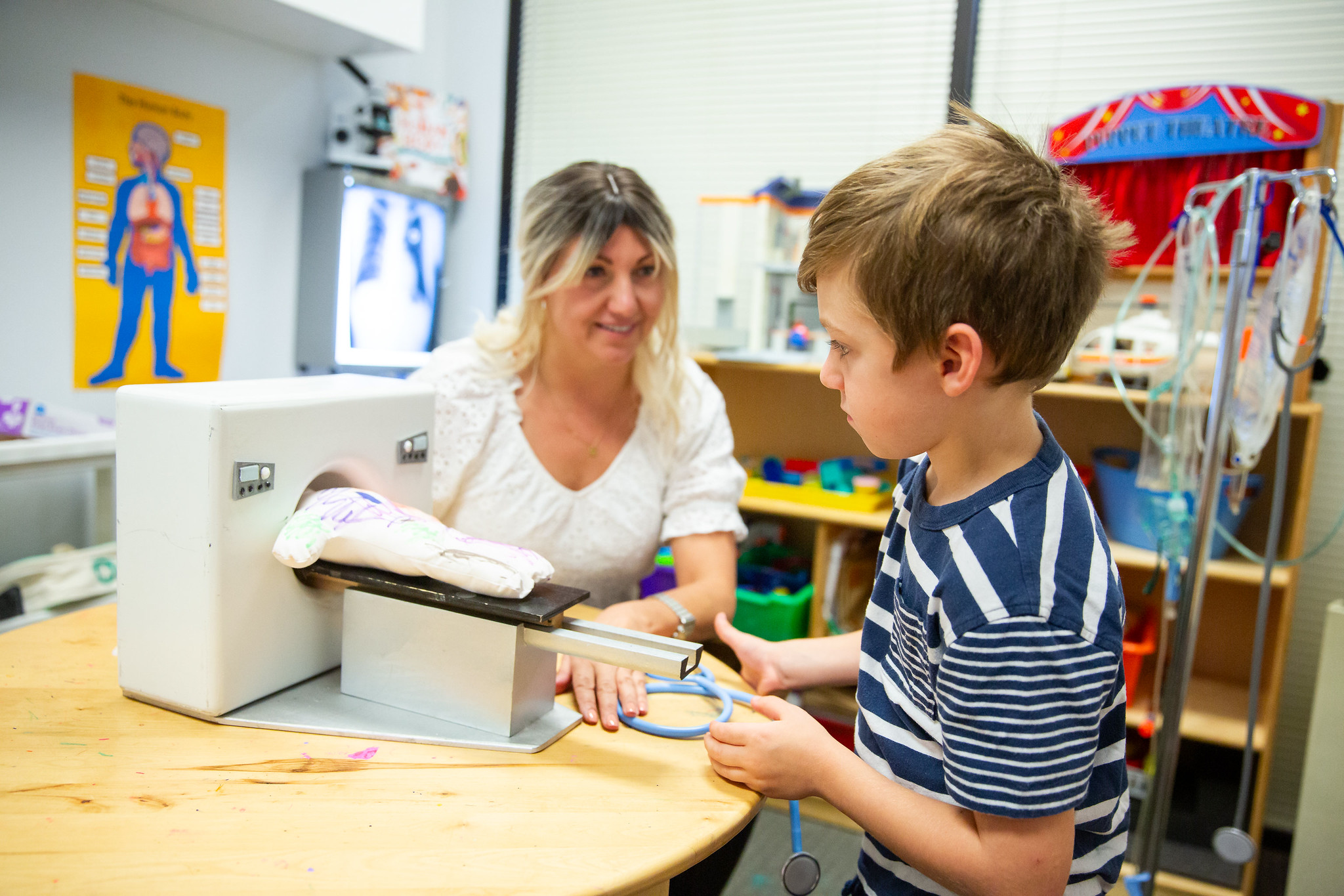 A woman and a boy interact at a medical or therapy setting, with colorful educational posters and equipment in the background.