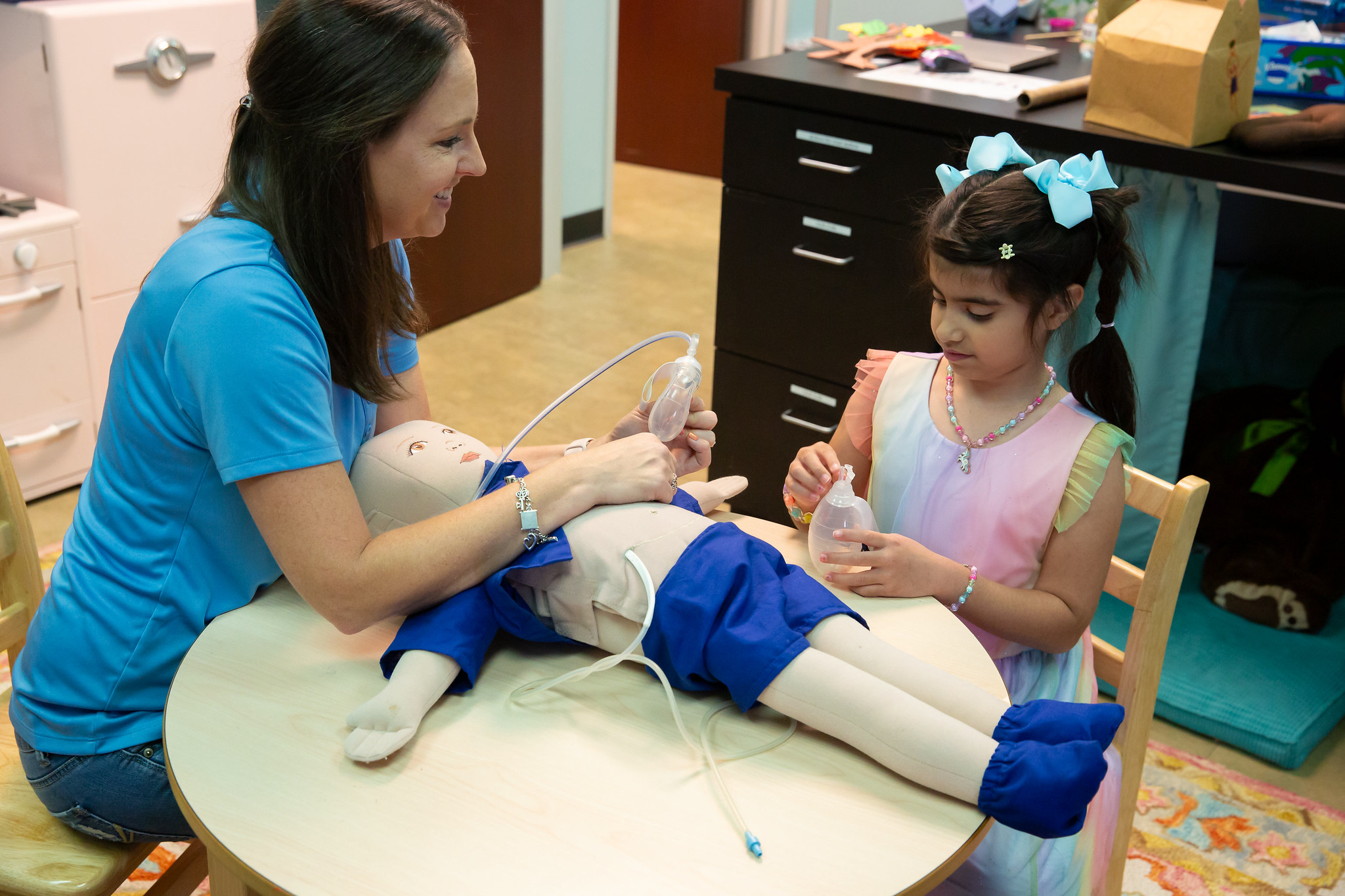 A nurse and a young girl with a bow in her hair are at a table with a medical mannequin, practicing medical procedures.