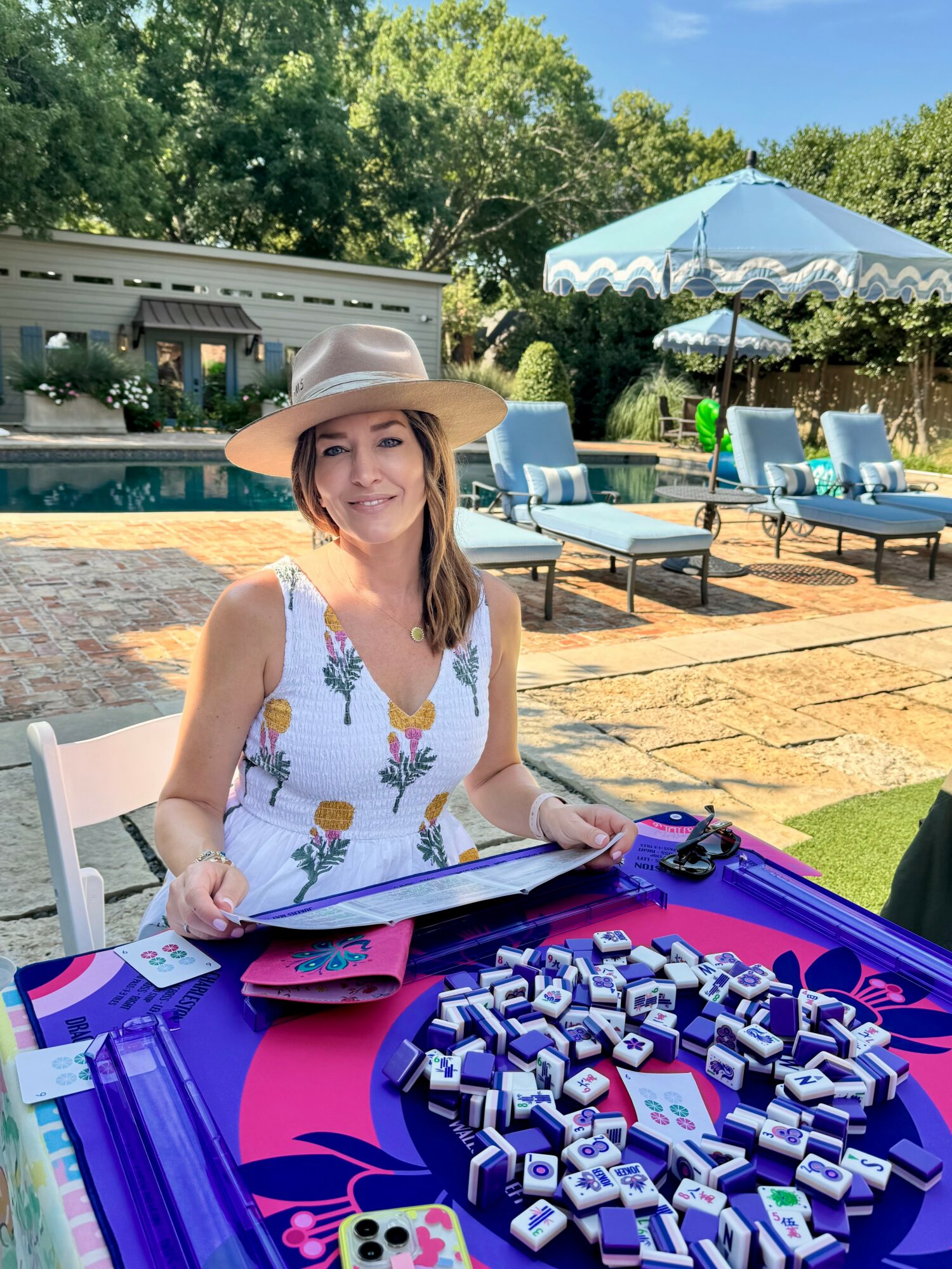 Woman sitting at table outdoors with pool, lounge chairs, and umbrella in background, smiling, with papers and game pieces on table.