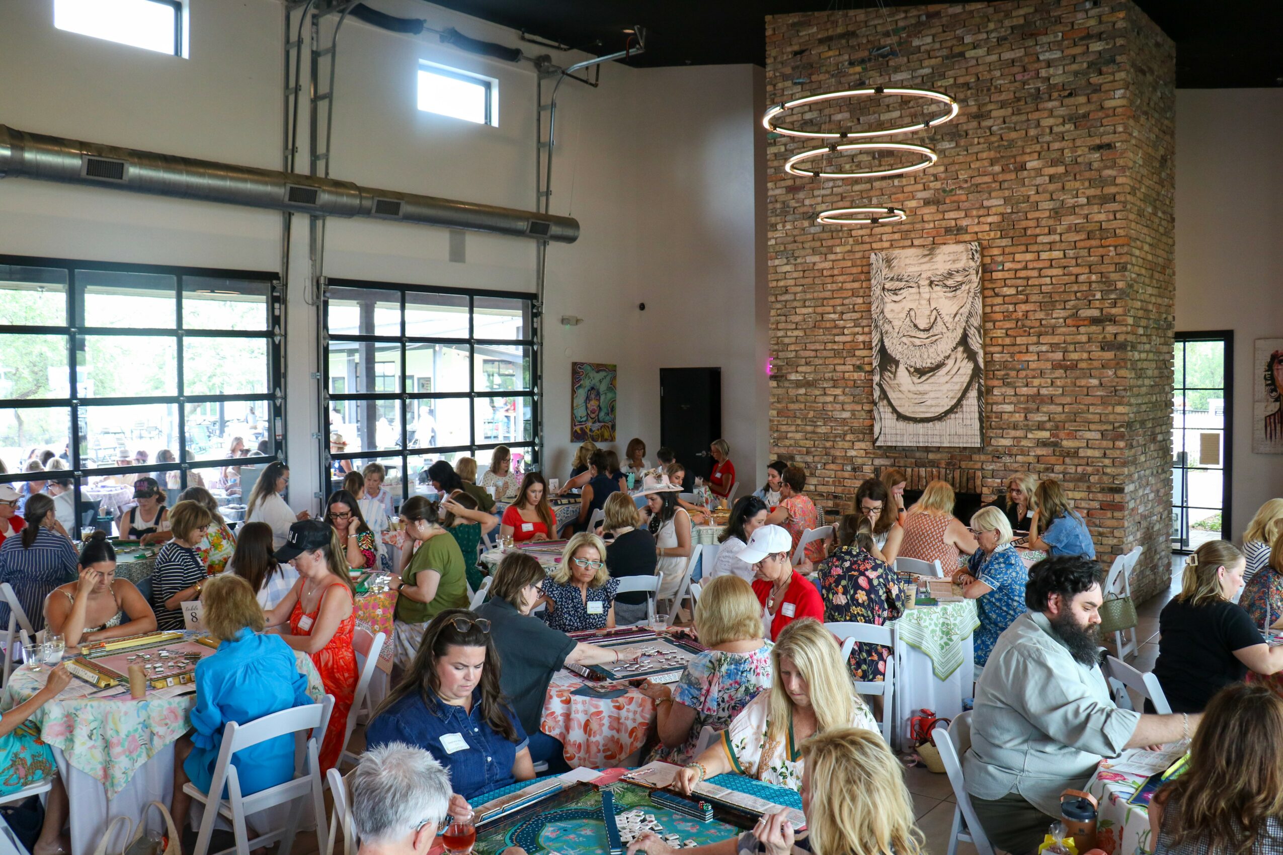 People seated at tables in a spacious, industrial-style room with large windows and a brick wall with artwork.