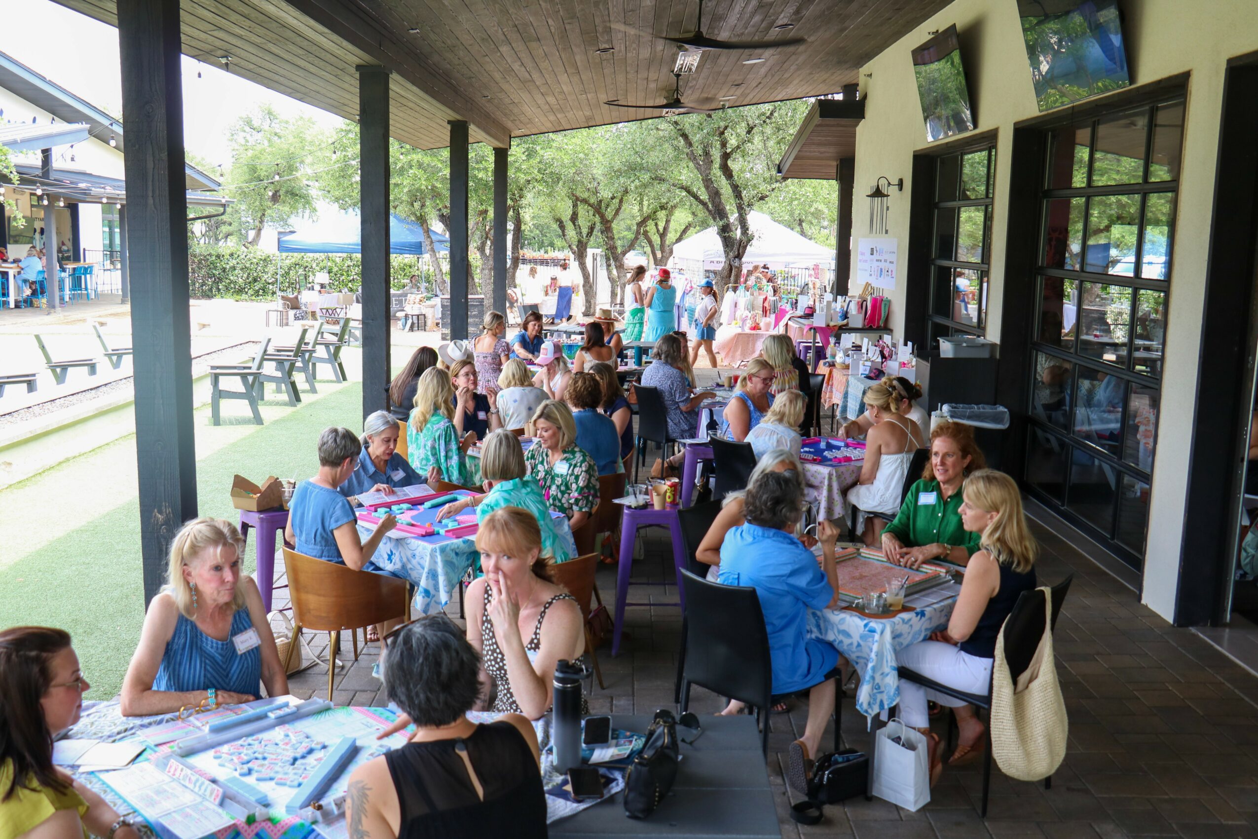 People dining at tables under a covered outdoor area with large windows, trees, and outdoor seating visible in the background.