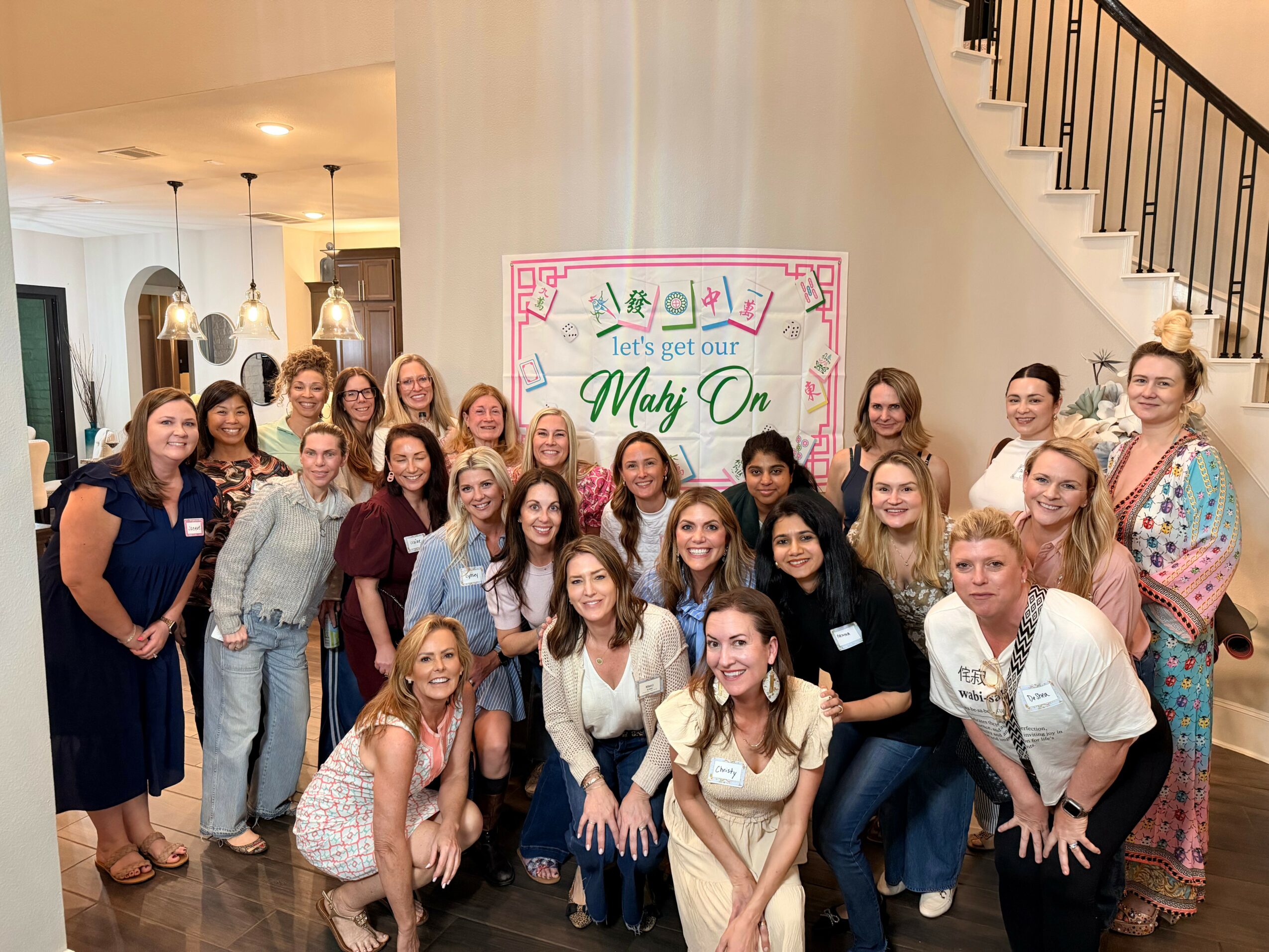 Group of women gathered indoors in front of a decorated sign, smiling for a photo, with a staircase in the background.