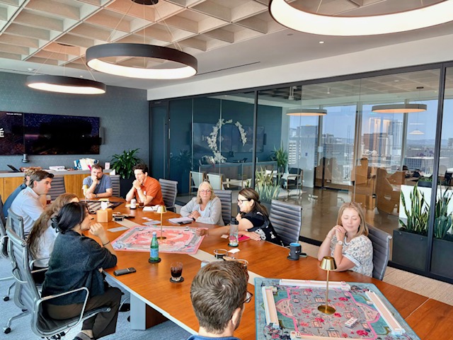 People sitting around a conference table in a modern office, with large windows and city view, discussing and working.