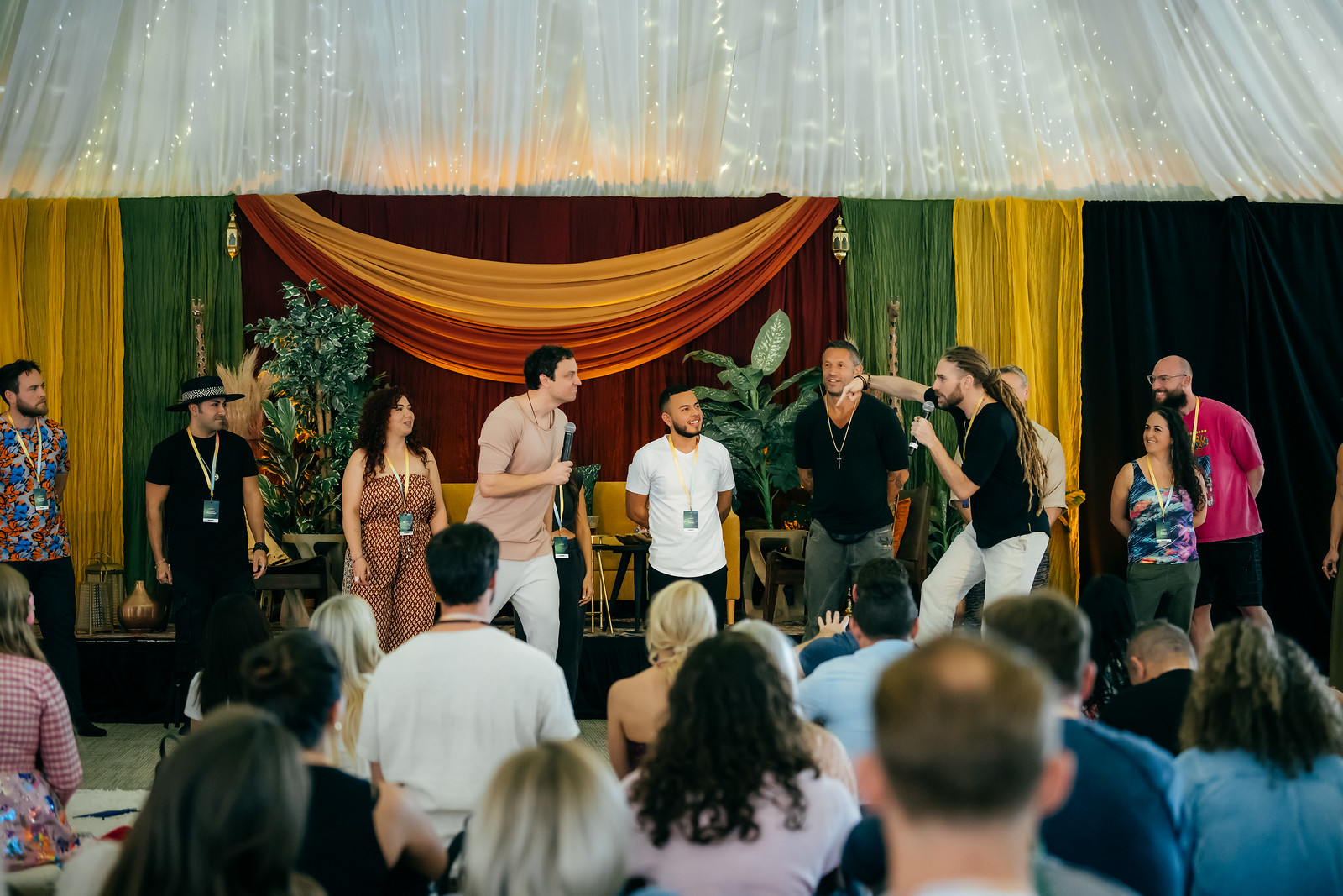 Group of people on stage with colorful curtains and plants, some speaking or performing, audience watching, indoor event.