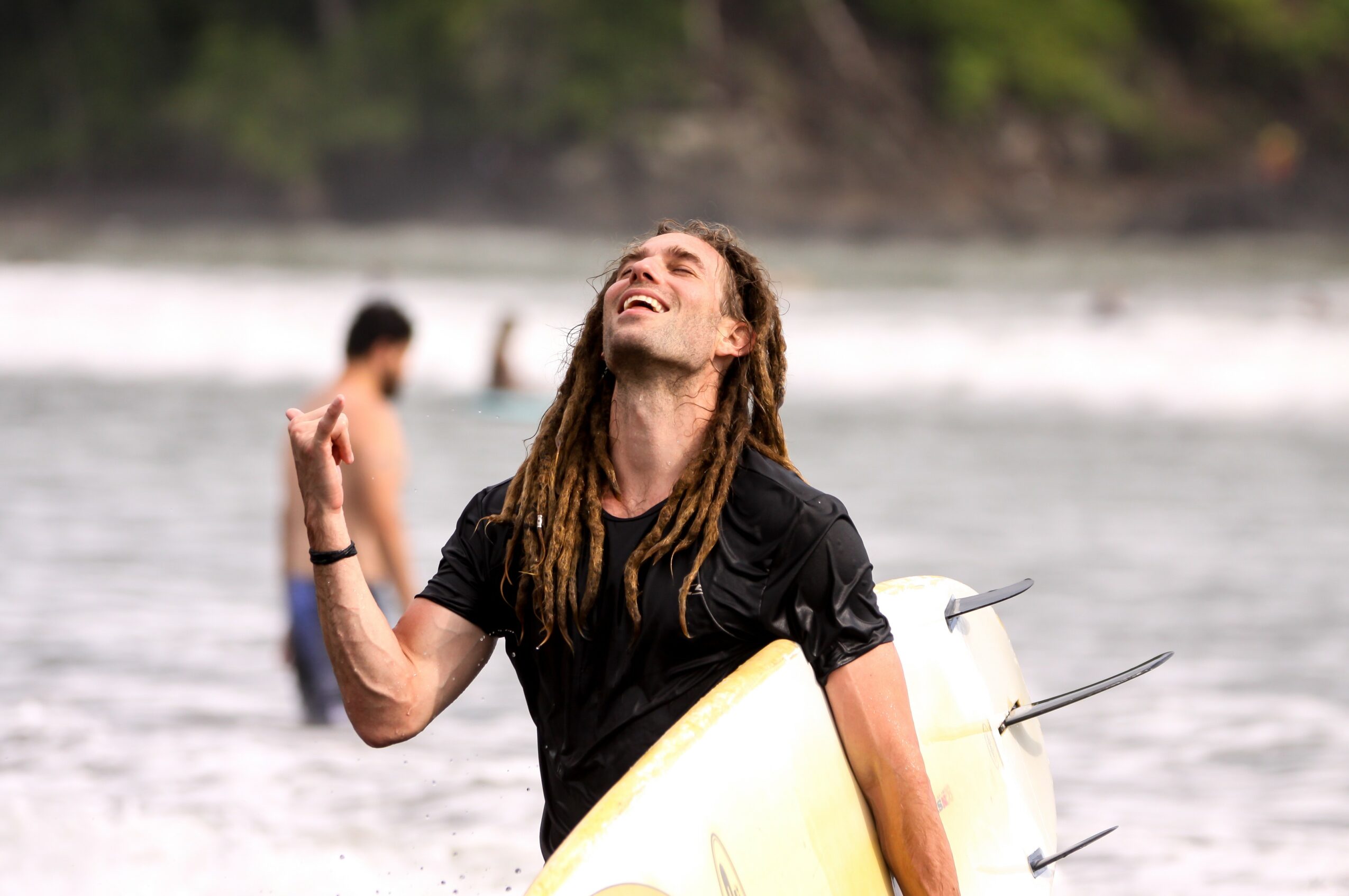 Person with long hair and a black shirt holding a surfboard on a beach, smiling and looking upward.