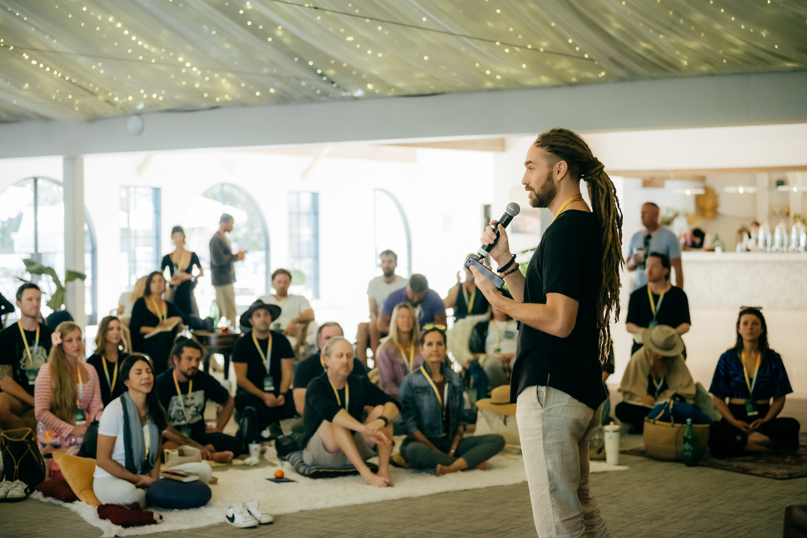 Man with long hair and beard speaking to seated audience in a bright, decorated indoor space, holding a microphone.