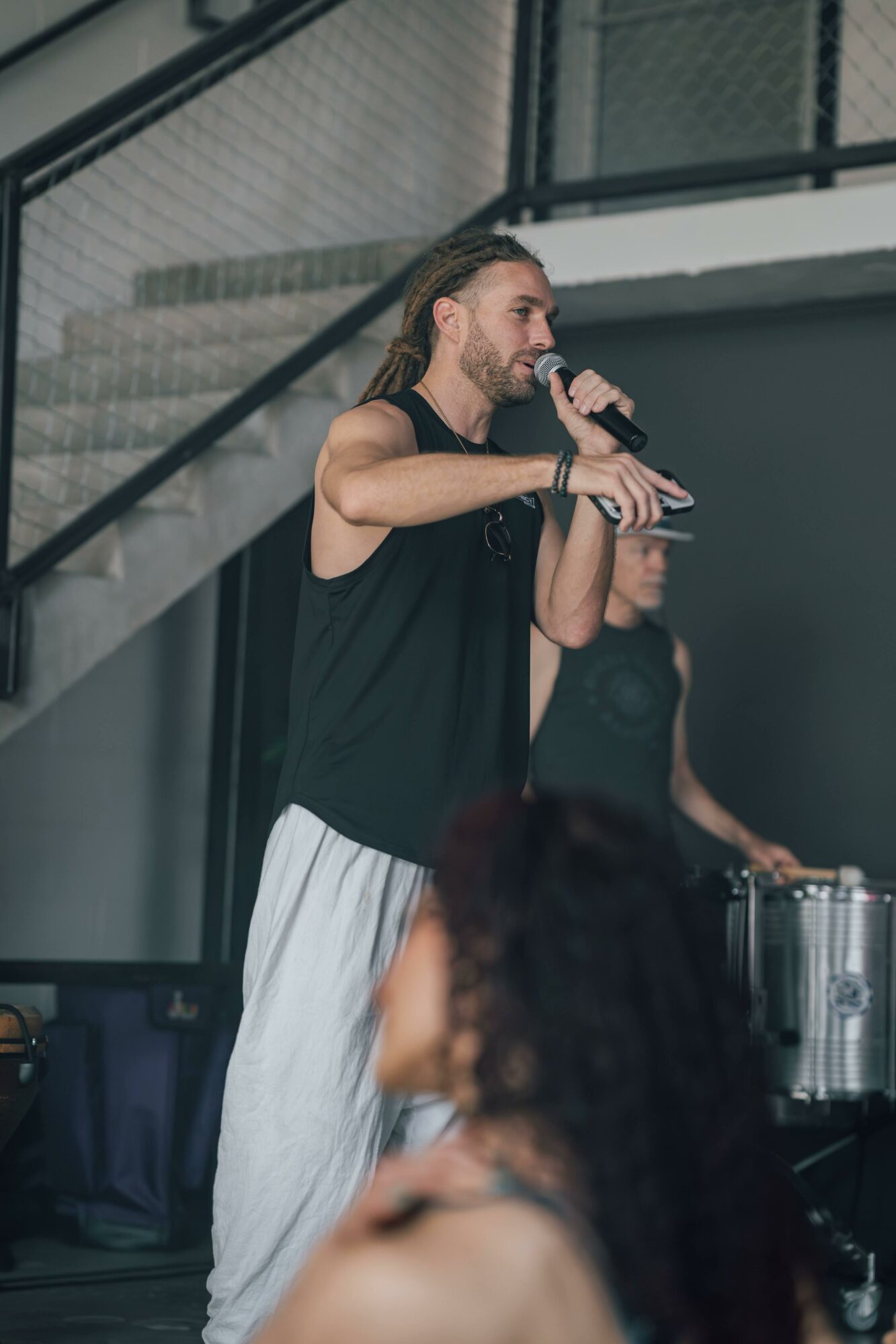 Person with long hair and beard speaking into a microphone, wearing a black sleeveless top and white pants, indoors.