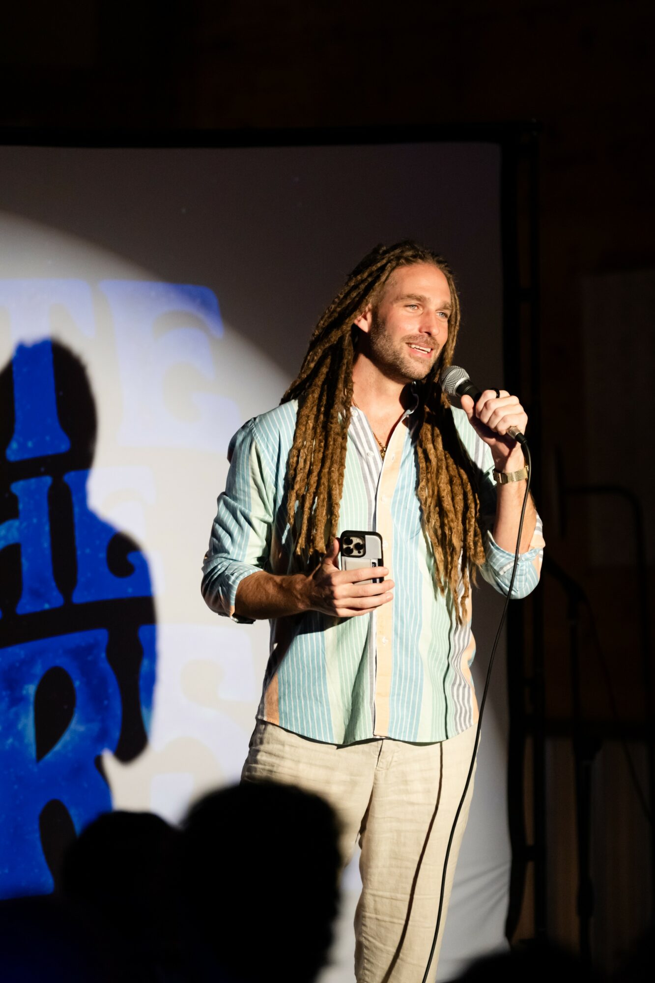 Person with long dreadlocks holding a microphone and a phone, standing on stage with a large screen behind.