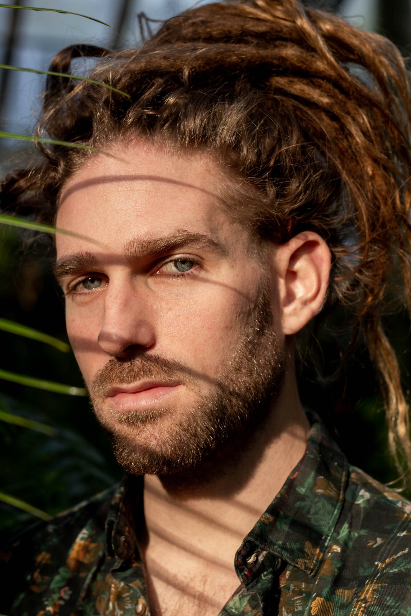 Close-up of a man with dreadlocks, beard, and intense gaze, outdoors with greenery in the background.
