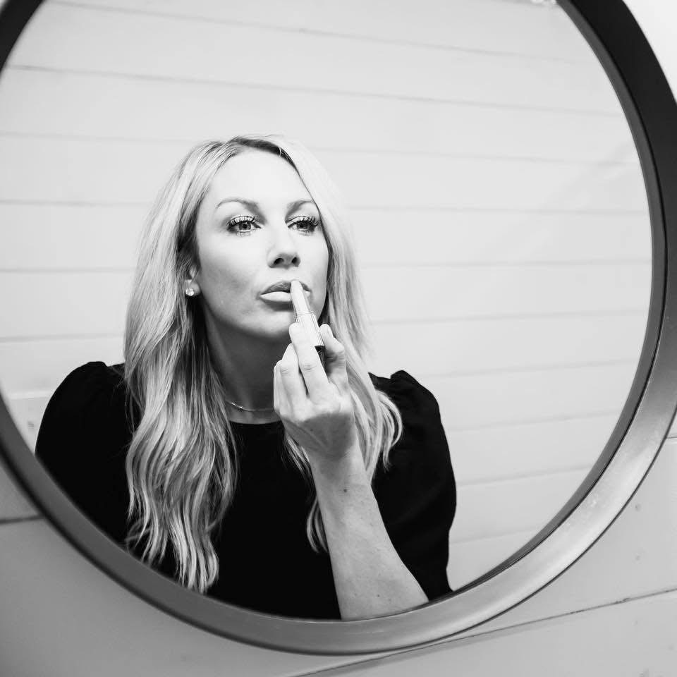 Woman looking into a mirror, touching her lips with her finger, with a neutral background.