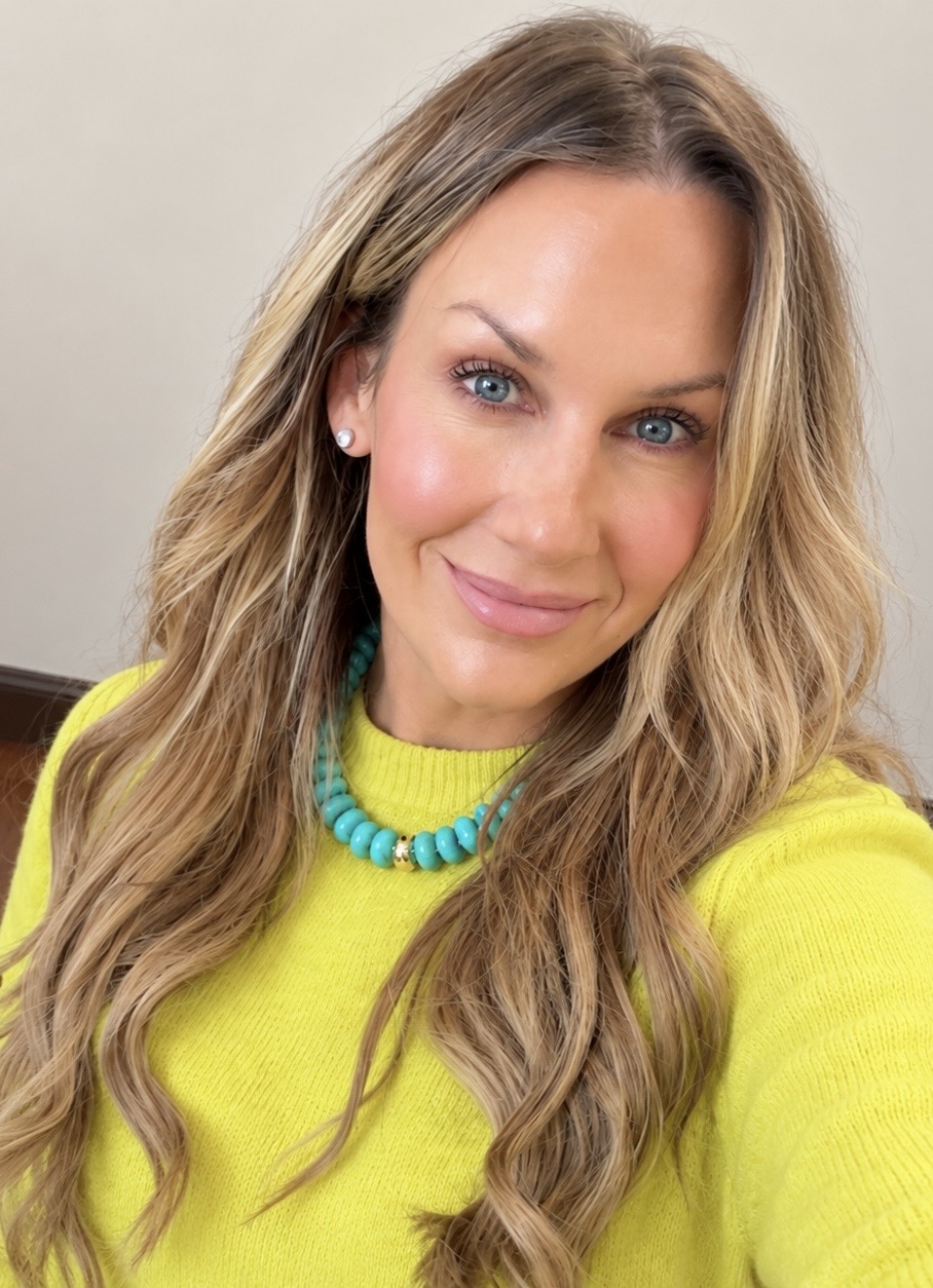 Woman with long wavy hair wearing a yellow sweater and turquoise necklace, smiling at the camera.