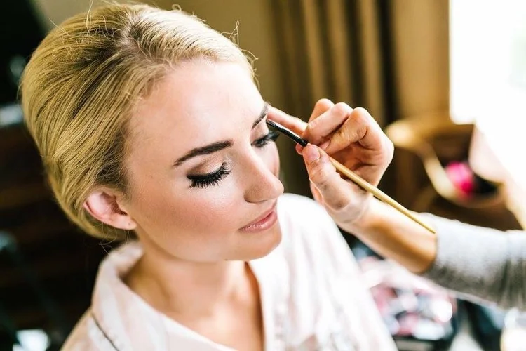 Woman getting makeup applied by a makeup artist, close-up of face and eye makeup, indoors with natural light.