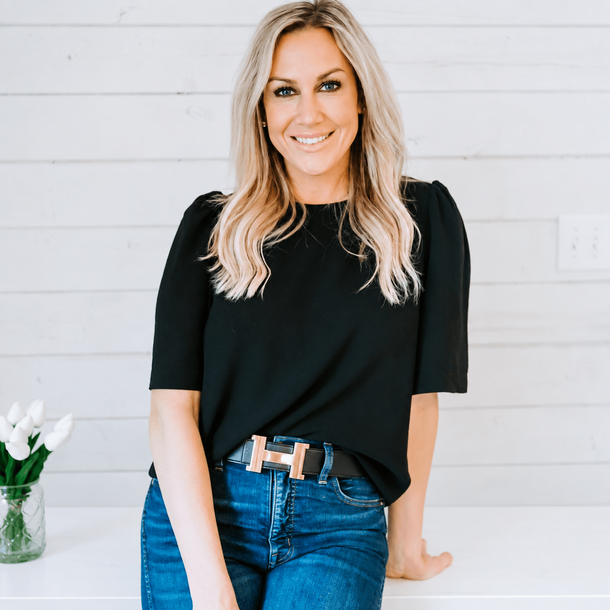 Smiling woman with blonde hair in a black top and blue jeans, standing against a white wooden wall, with a flower vase nearby.