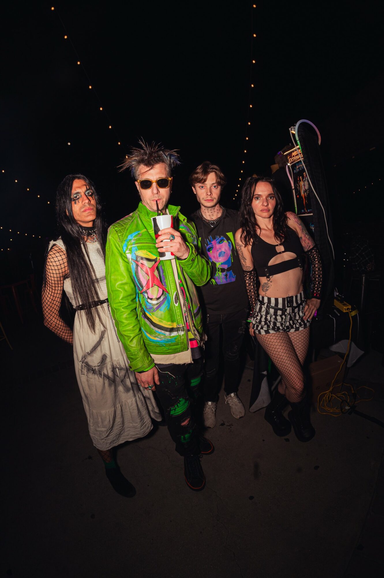 Group of five people standing together in a dark setting with string lights overhead, some holding musical instruments.