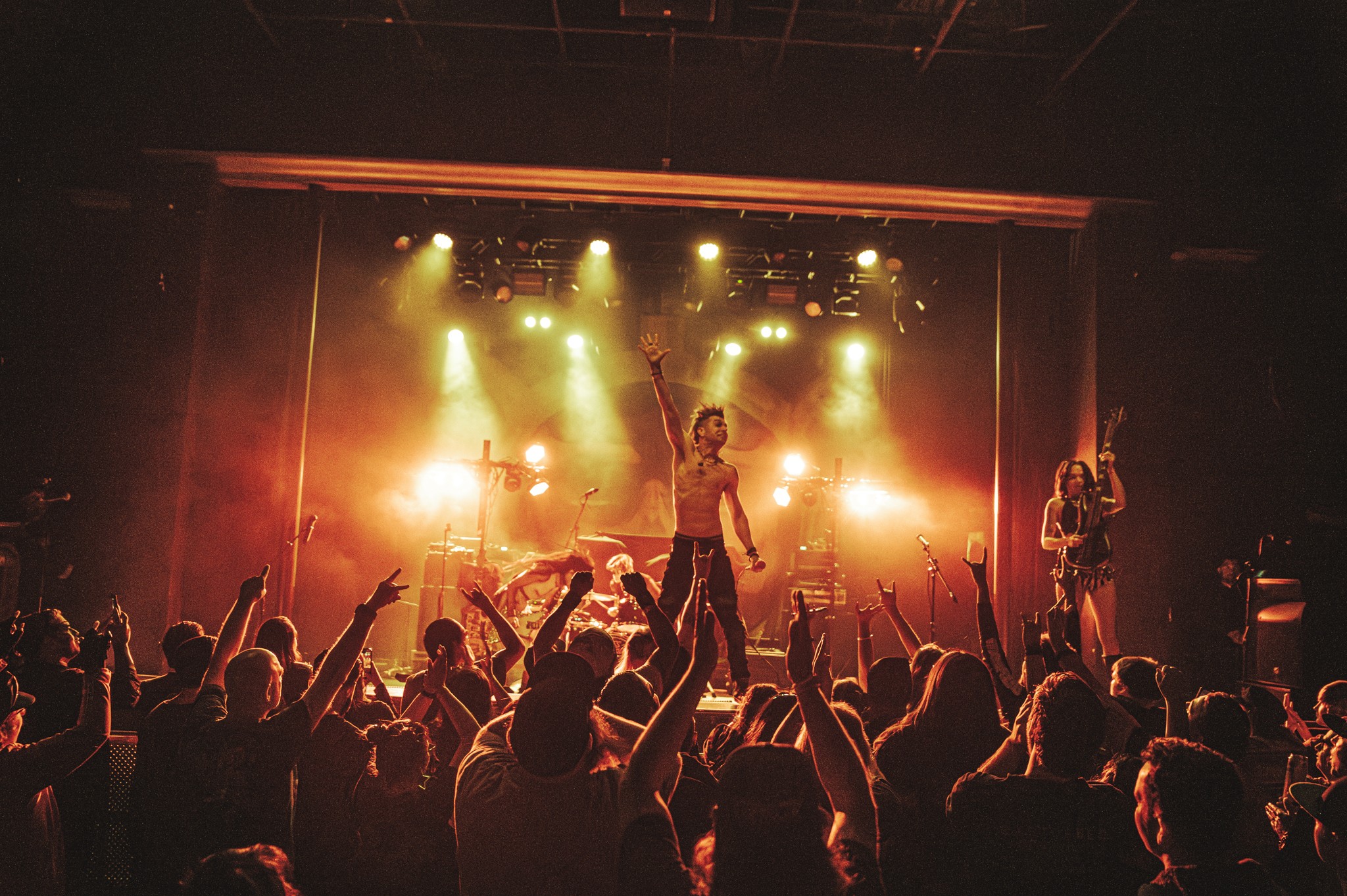 Concert scene with a band performing on stage, bright lights, and an audience with raised hands.