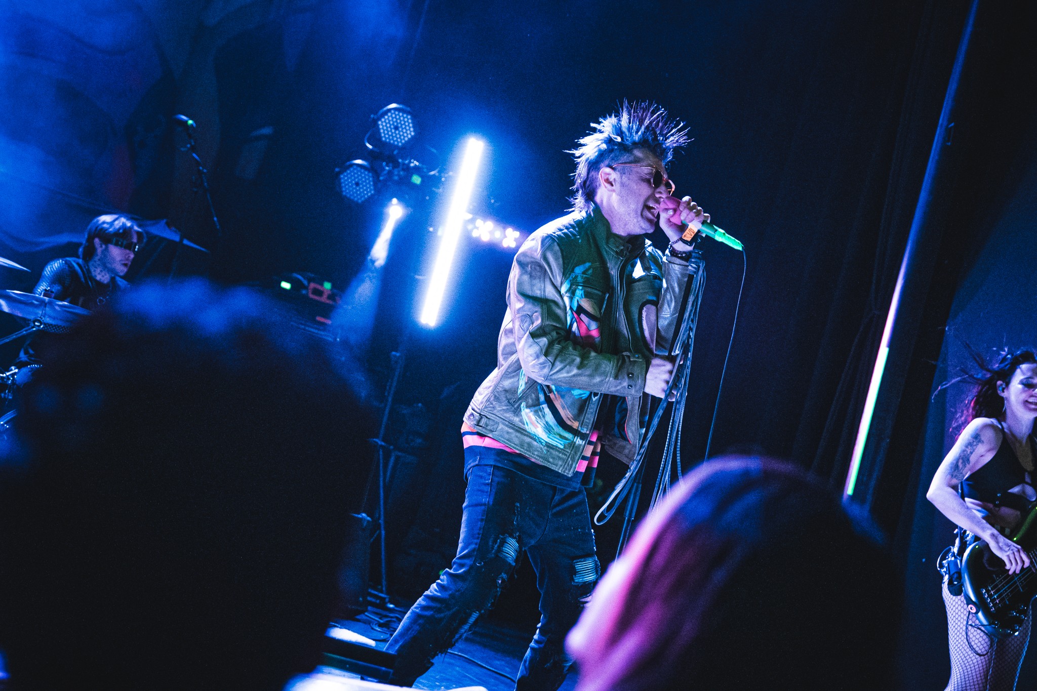 Performer with spiky hair singing into a microphone on stage with blue lighting, surrounded by band members and audience.