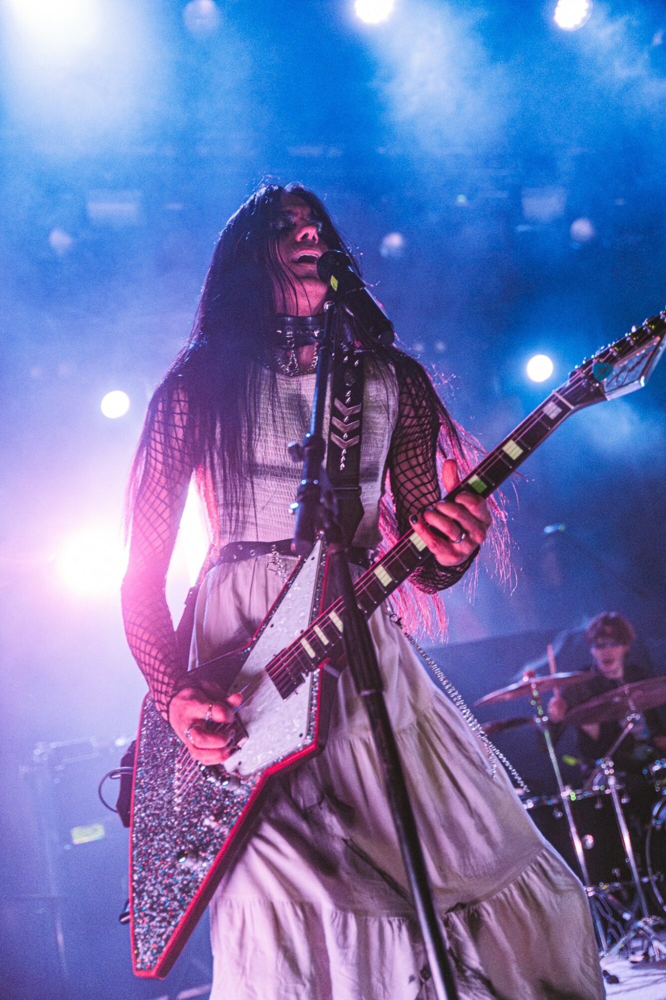 Female musician playing an electric guitar on stage with blue lighting, wearing a long dress and mesh top.