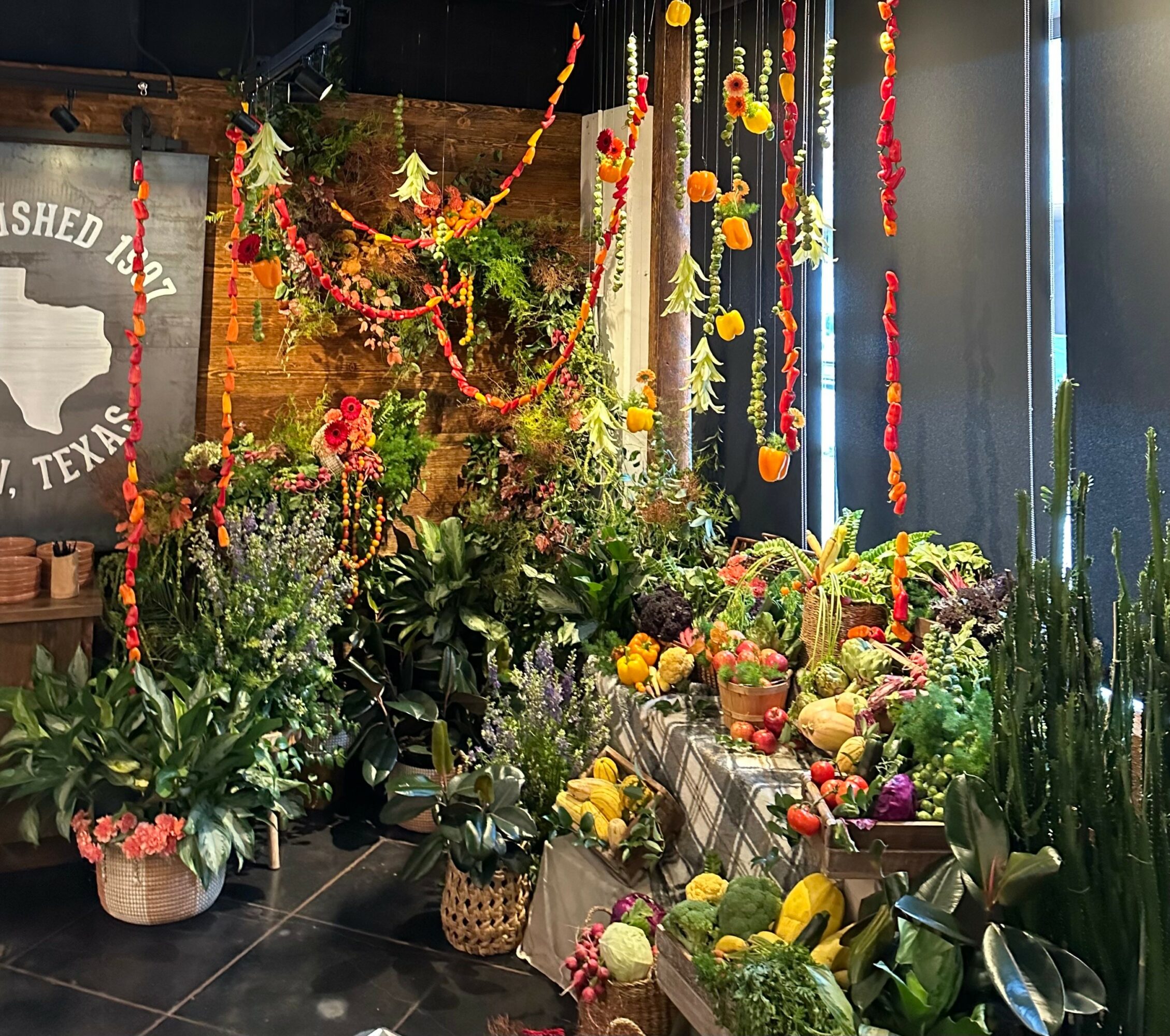 Display of colorful flowers, fruits, and vegetables with hanging decorations in a room, with a Texas-themed sign on the wall.