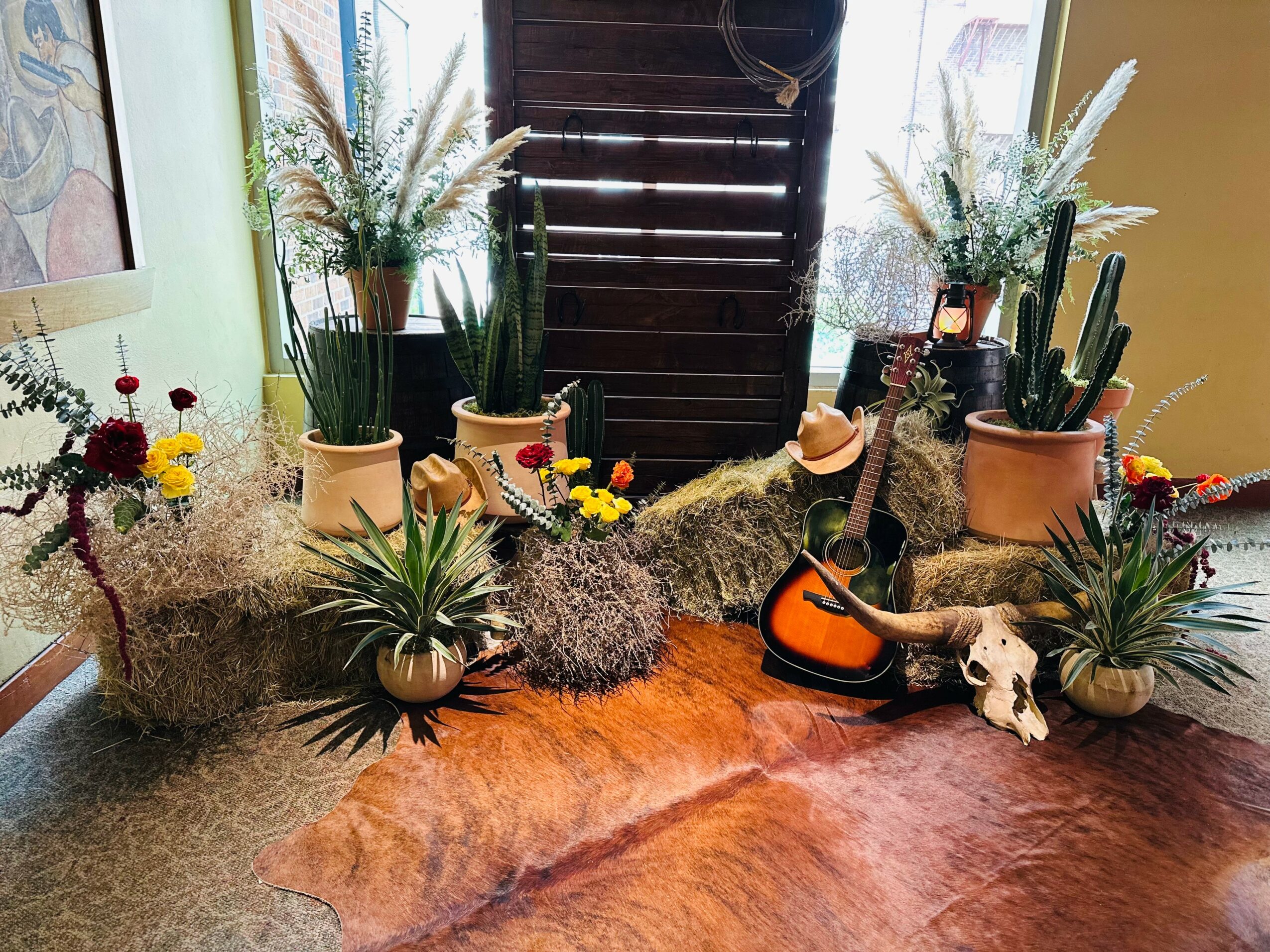Decorative arrangement of potted plants, flowers, and a small animal skull on a wooden table, with a window and blinds in the background.